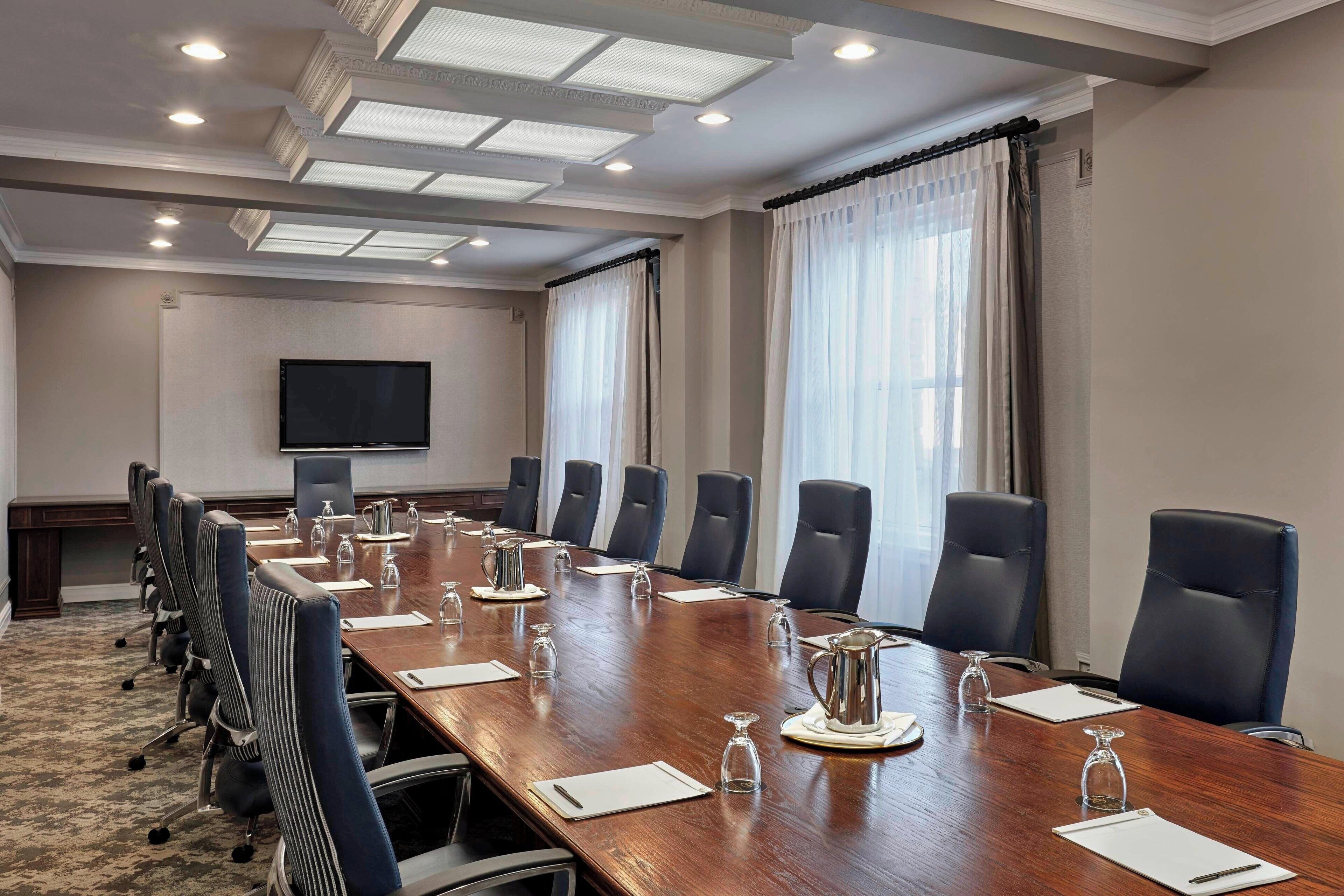 Un salón de conferencias con una larga mesa, varias sillas, una televisión y grandes ventanales con cortinas.