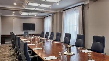 Un salón de conferencias con una larga mesa, varias sillas, una televisión y grandes ventanales con cortinas.