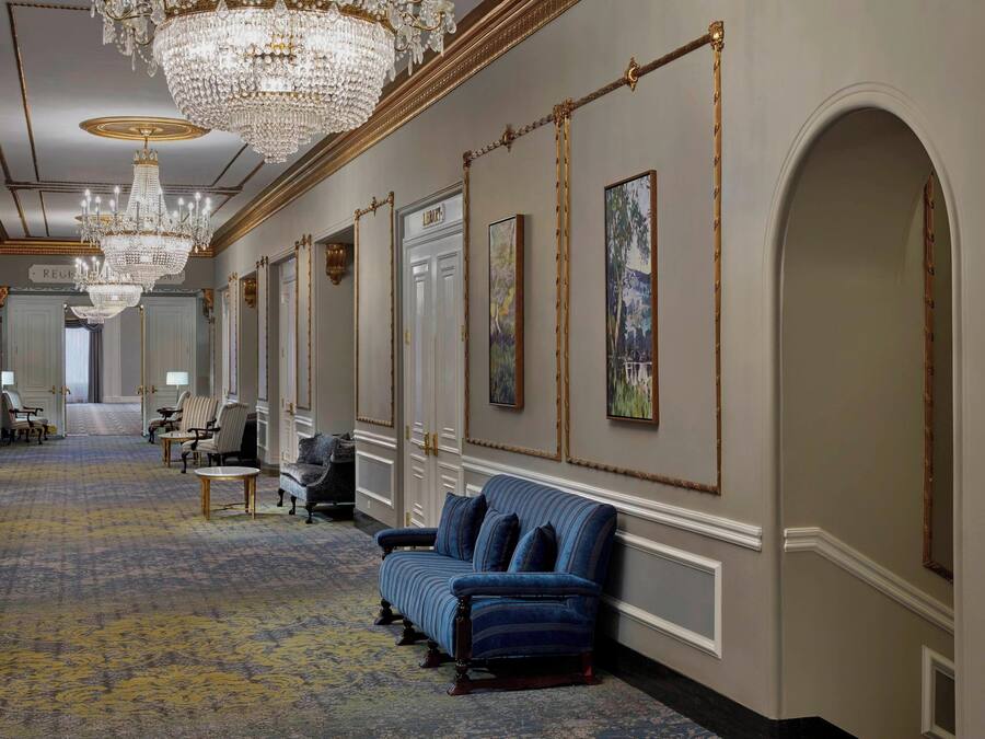 A grand hallway with chandeliers, a blue sofa, and framed artwork on the walls.