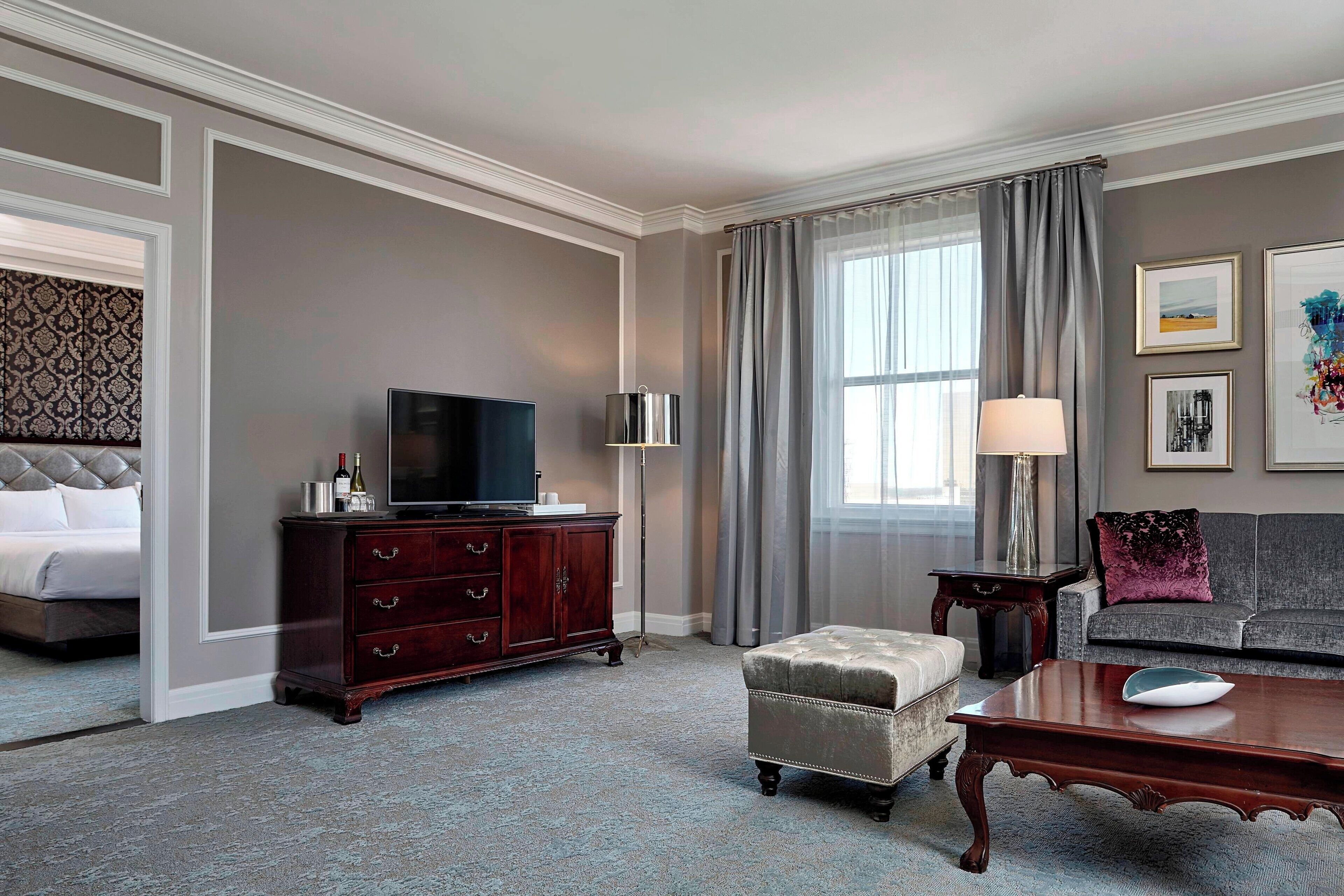 Habitación de hotel con cama, televisor, cómoda de madera, sofá, mesa de centro y ventana con cortinas.
