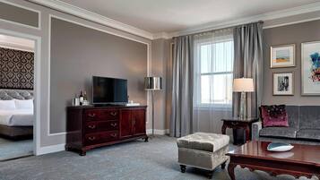 Habitación de hotel con cama, televisor, cómoda de madera, sofá, mesa de centro y ventana con cortinas.