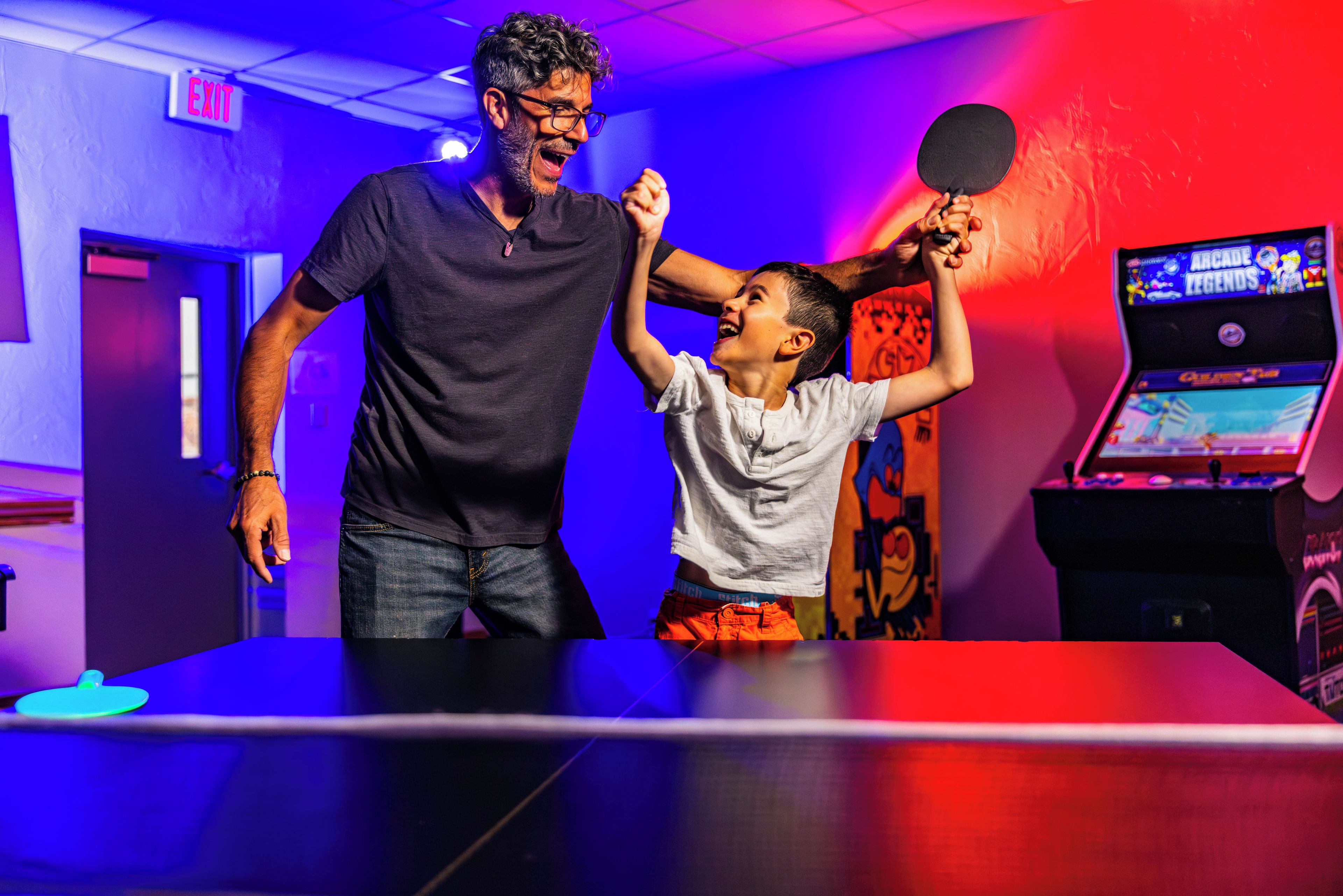 Un homme et un enfant jouent au ping-pong dans une pièce où l’on peut voir un panneau de sortie lumineux et une borne d’arcade.