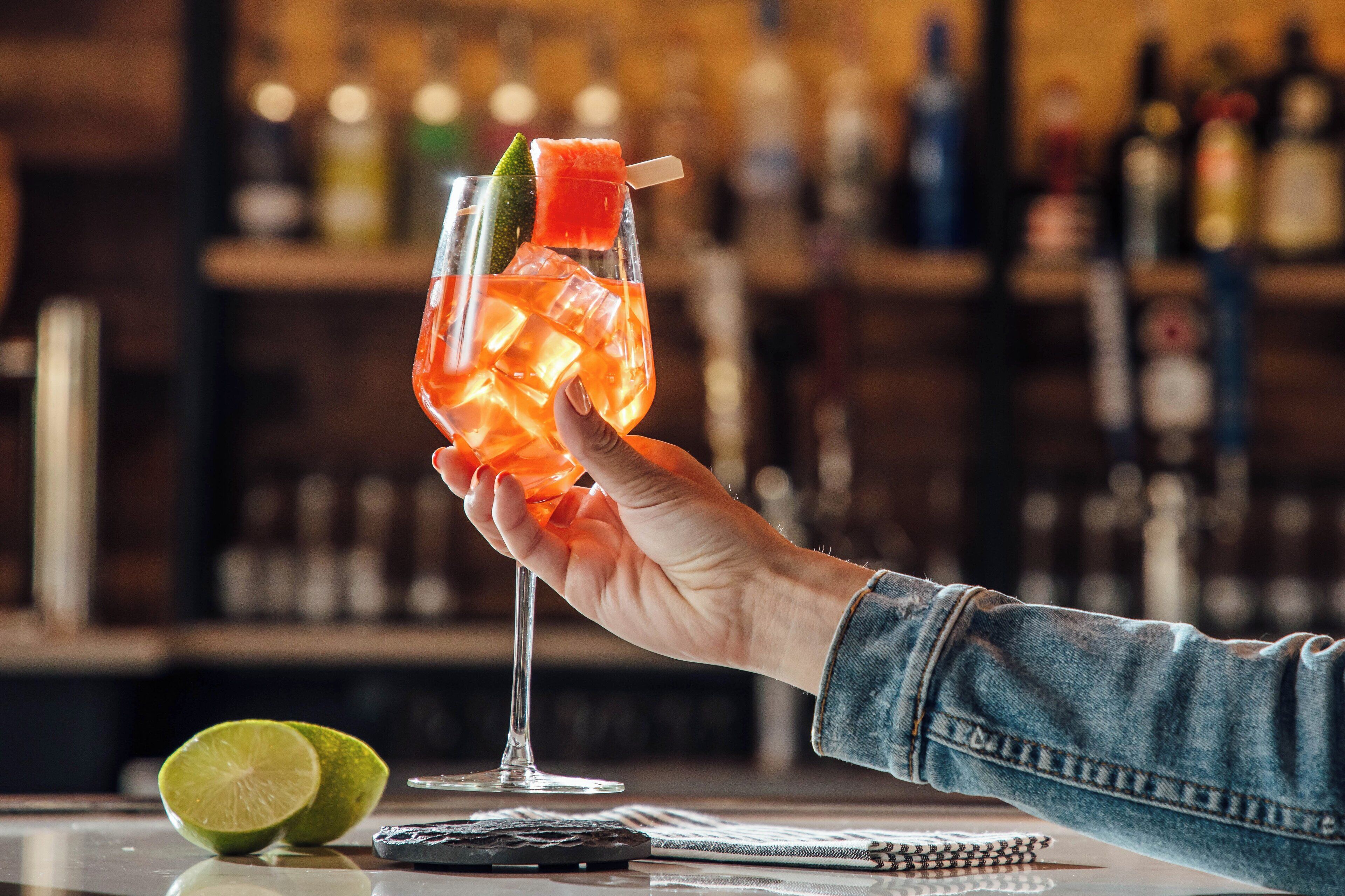 A hand holding a cocktail with ice cubes and a lime wedge, with a blurred bar background.