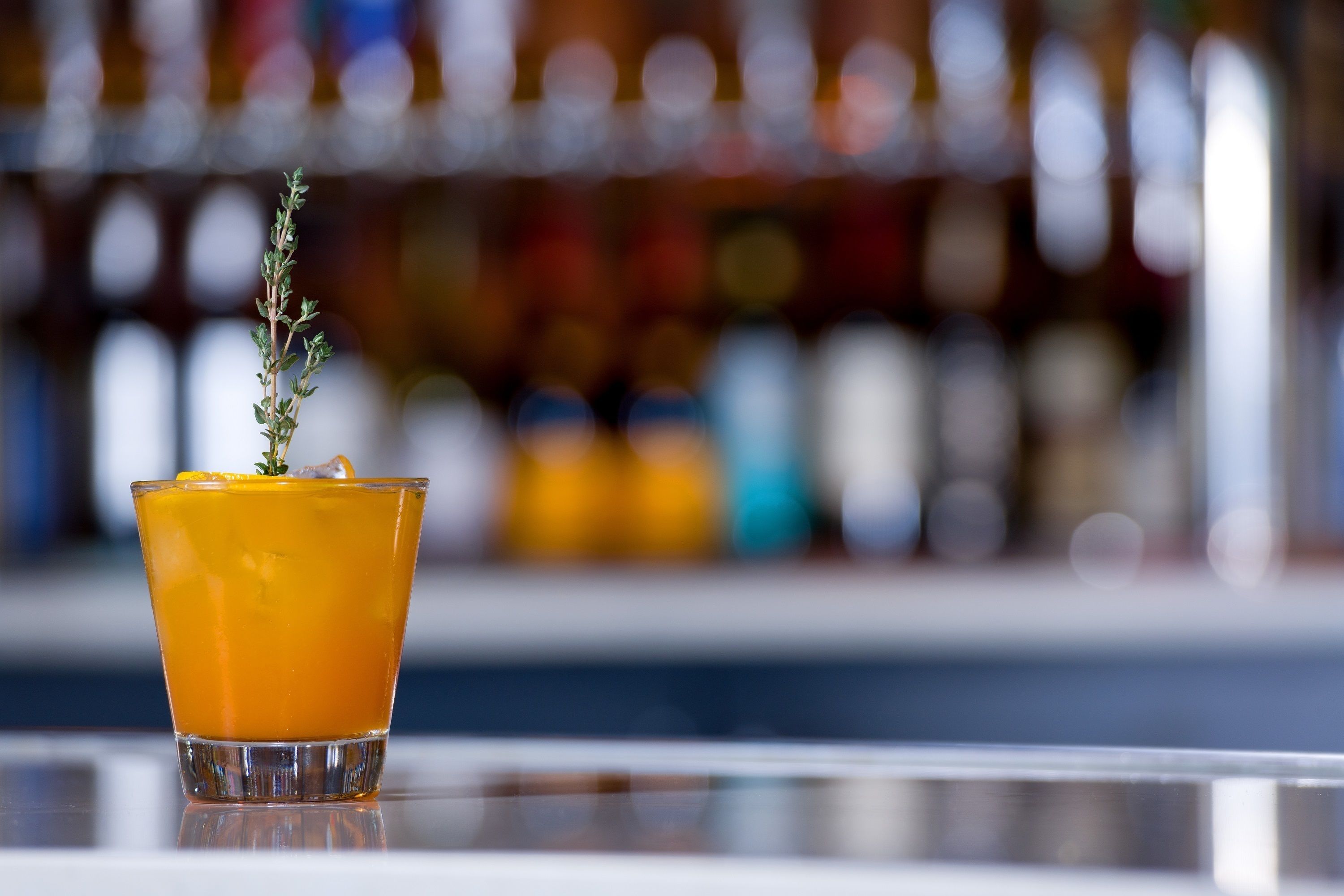 A glass of orange drink with a sprig of thyme on the rim, placed on a reflective surface with a blurred background of bottles.