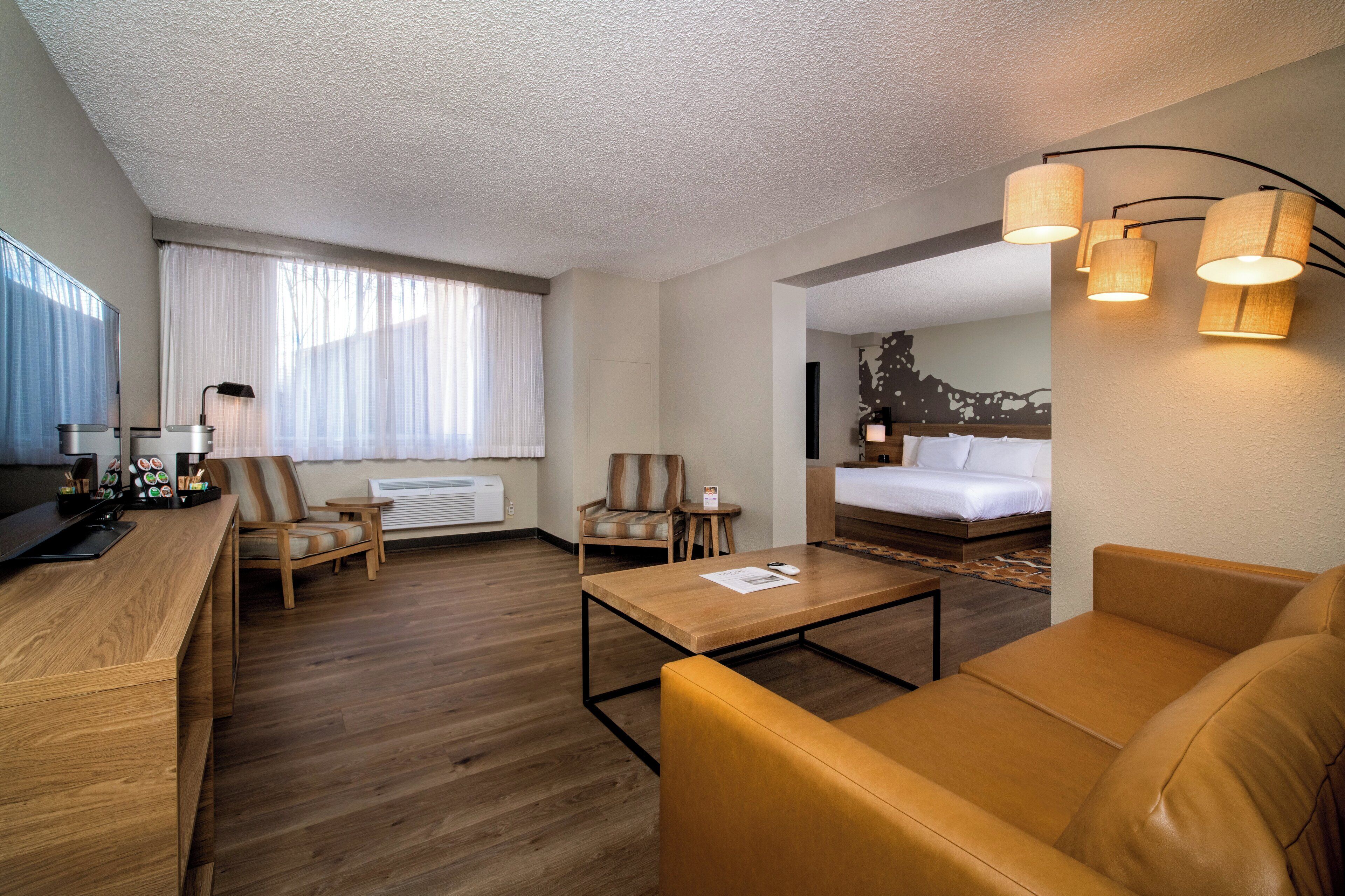A hotel room with a wooden desk, two armchairs, a sofa, a bed, and a large window with sheer curtains.