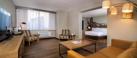 A hotel room with a wooden desk, two armchairs, a sofa, a bed, and a large window with sheer curtains.