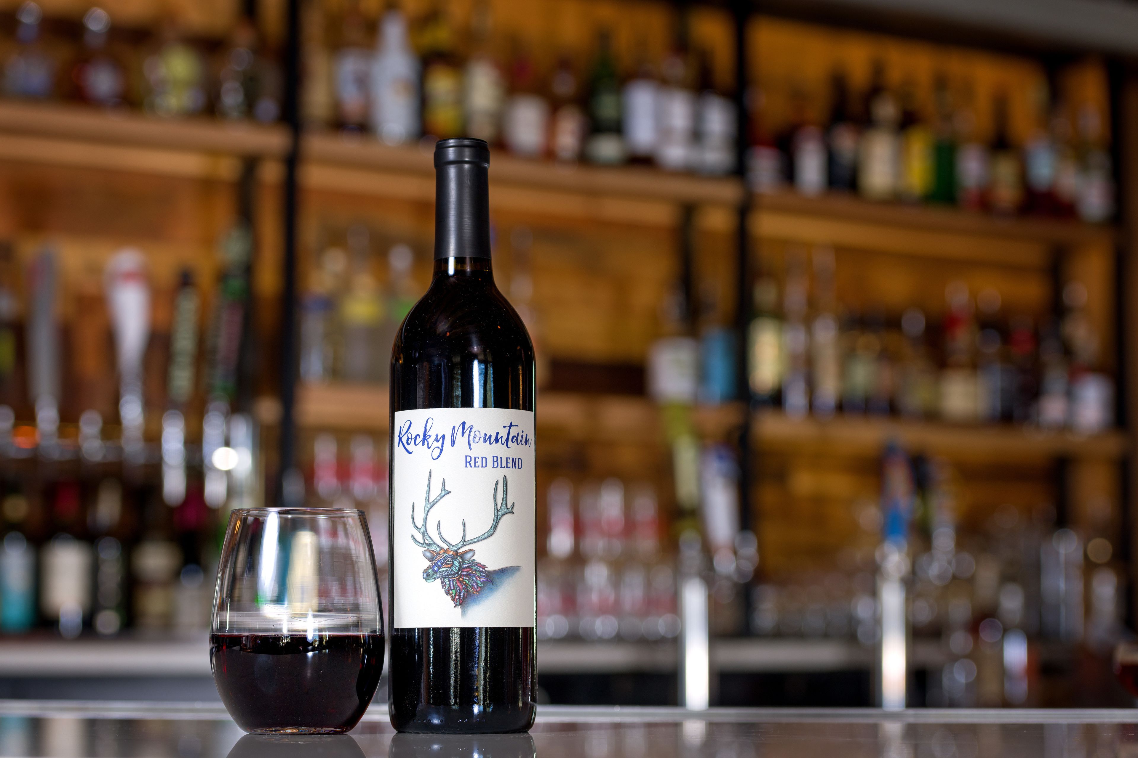 A bottle of Rocky Mountain Red Blend wine and a glass of red wine on a bar counter with shelves of various bottles in the background.
