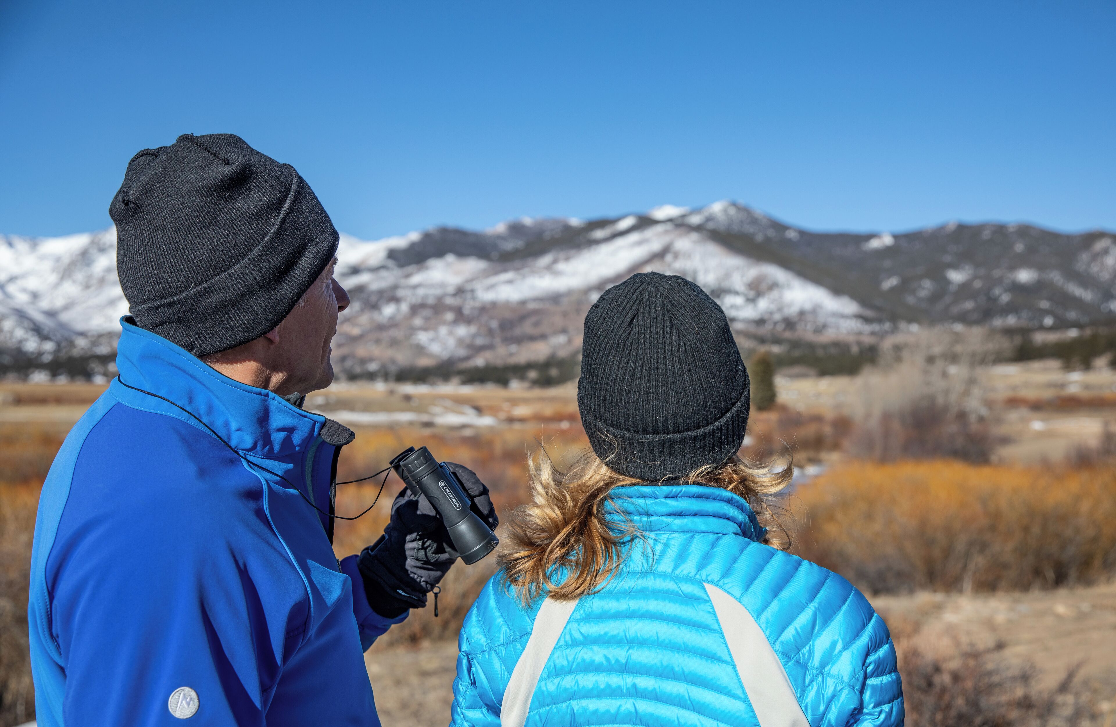 Due persone con giacche e berretti blu, una delle quali tiene in mano una macchina fotografica, in un paesaggio montano.