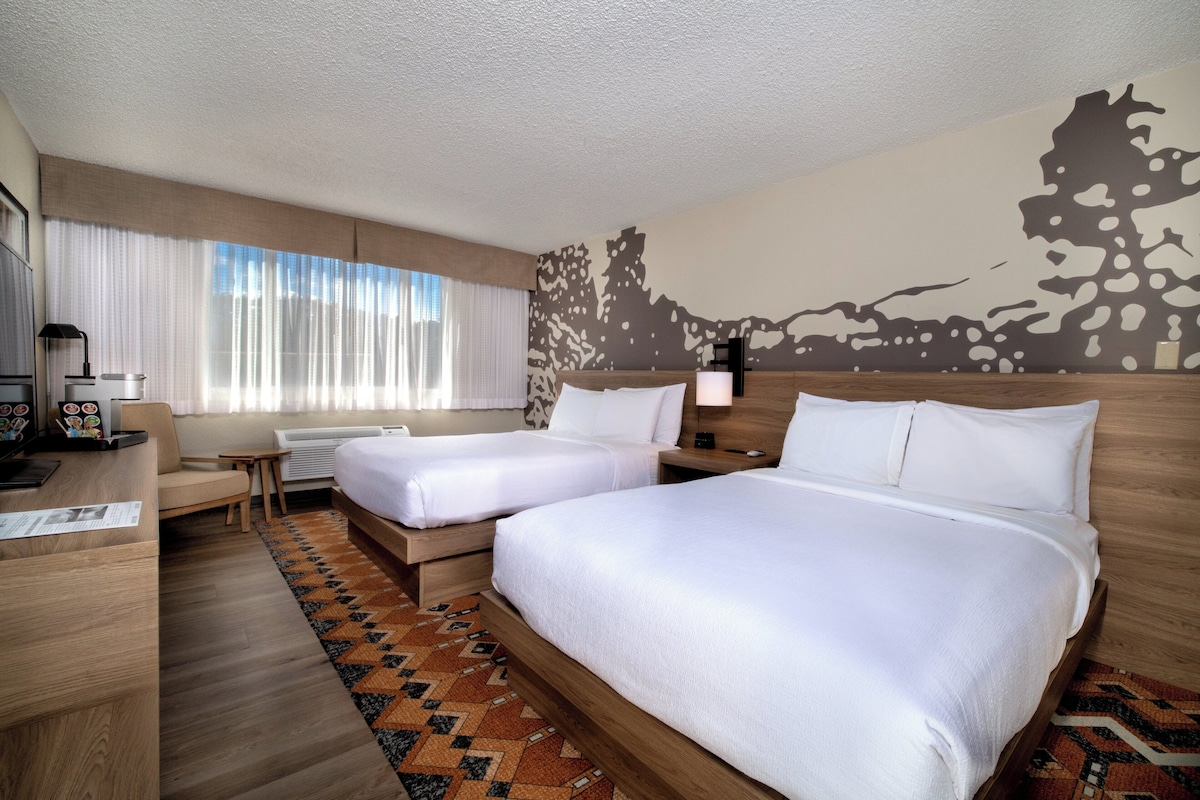 Une chambre d’hôtel avec deux lits, un bureau, une chaise et une grande fenêtre avec des rideaux légers.