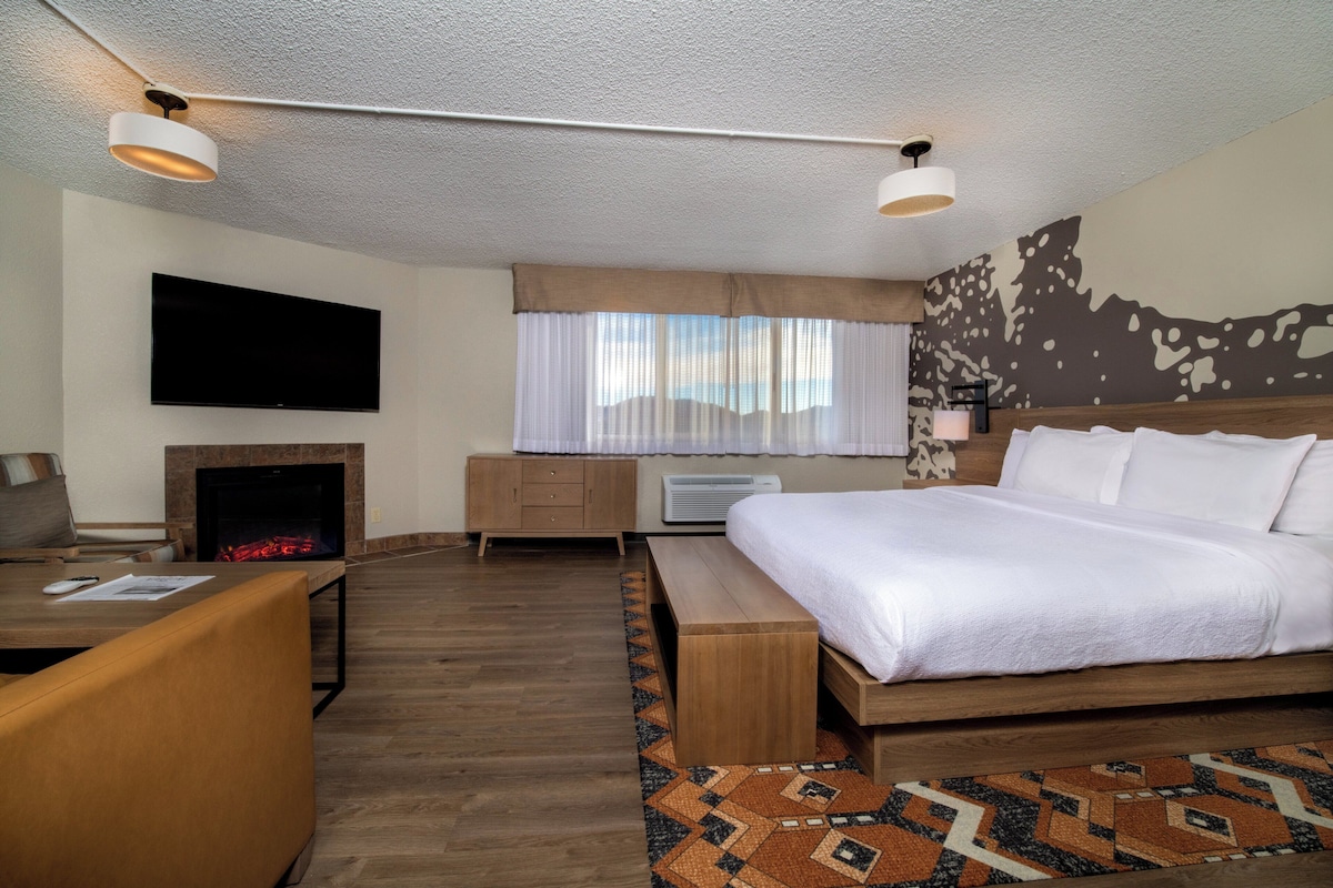 Une chambre d’hôtel moderne dotée d’un grand lit, d’une télévision à écran plat, d’une cheminée, d’un bureau et d’un tapis à motifs.