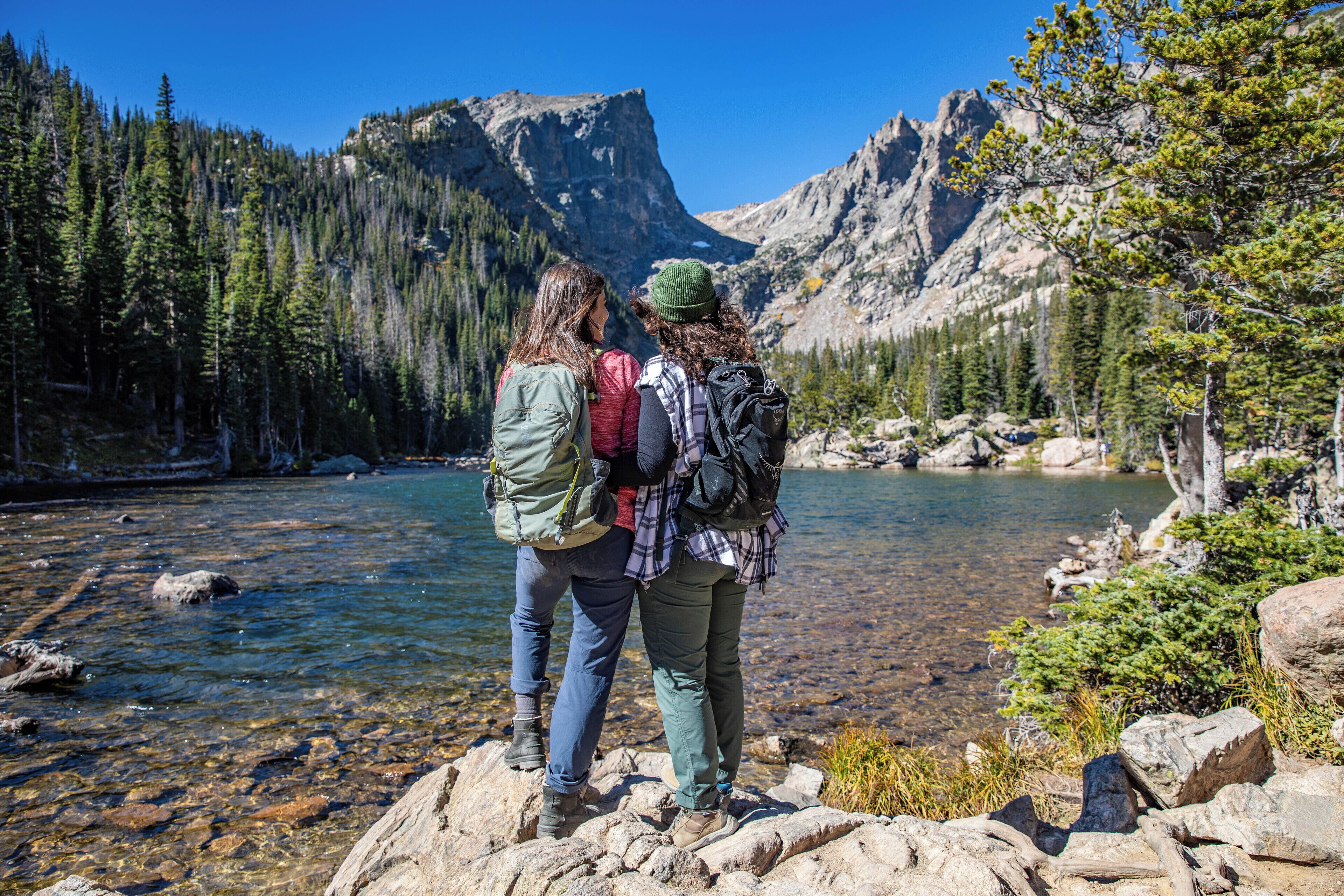 Two people standing by a clear mountain lake with rocky terrain and dense forest in the background.