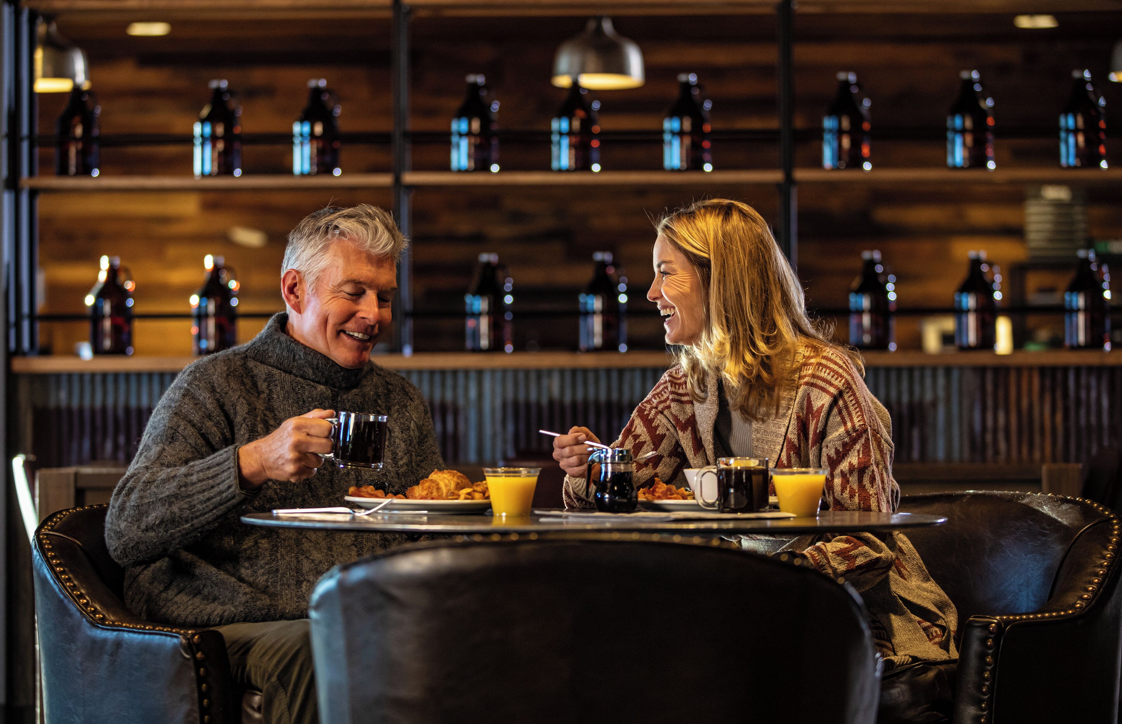Two people sitting at a table with breakfast items, in a room with shelves of bottles in the background.
