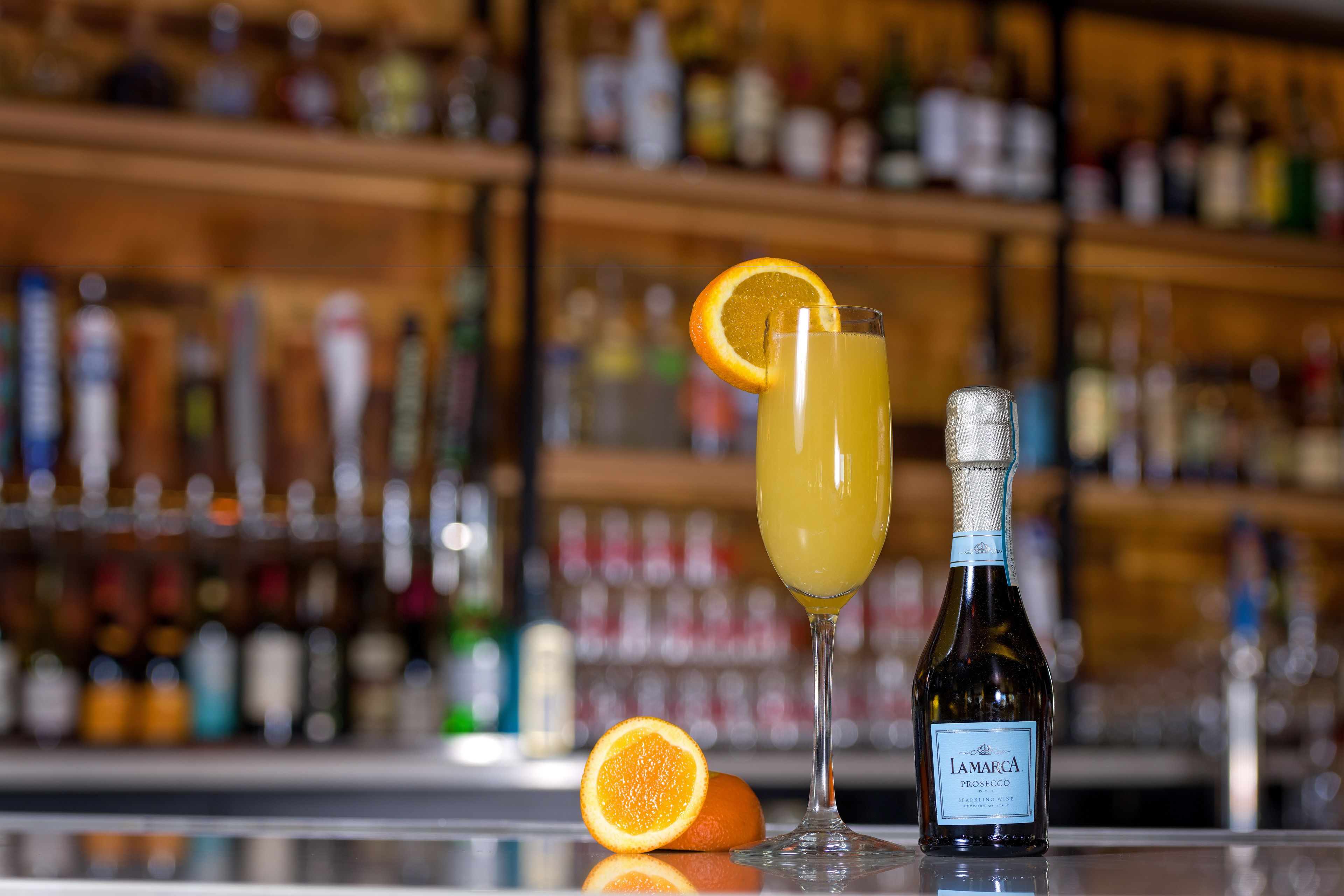 Un ambiente da bar con una bottiglia di Amaraca Prosecco e un bicchiere di cocktail guarnito con una fetta d'arancia.