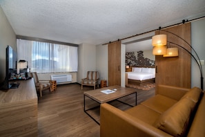 A hotel room with a wooden floor, a sofa, a desk with a lamp, a chair, a bed, and a large window with sheer curtains.