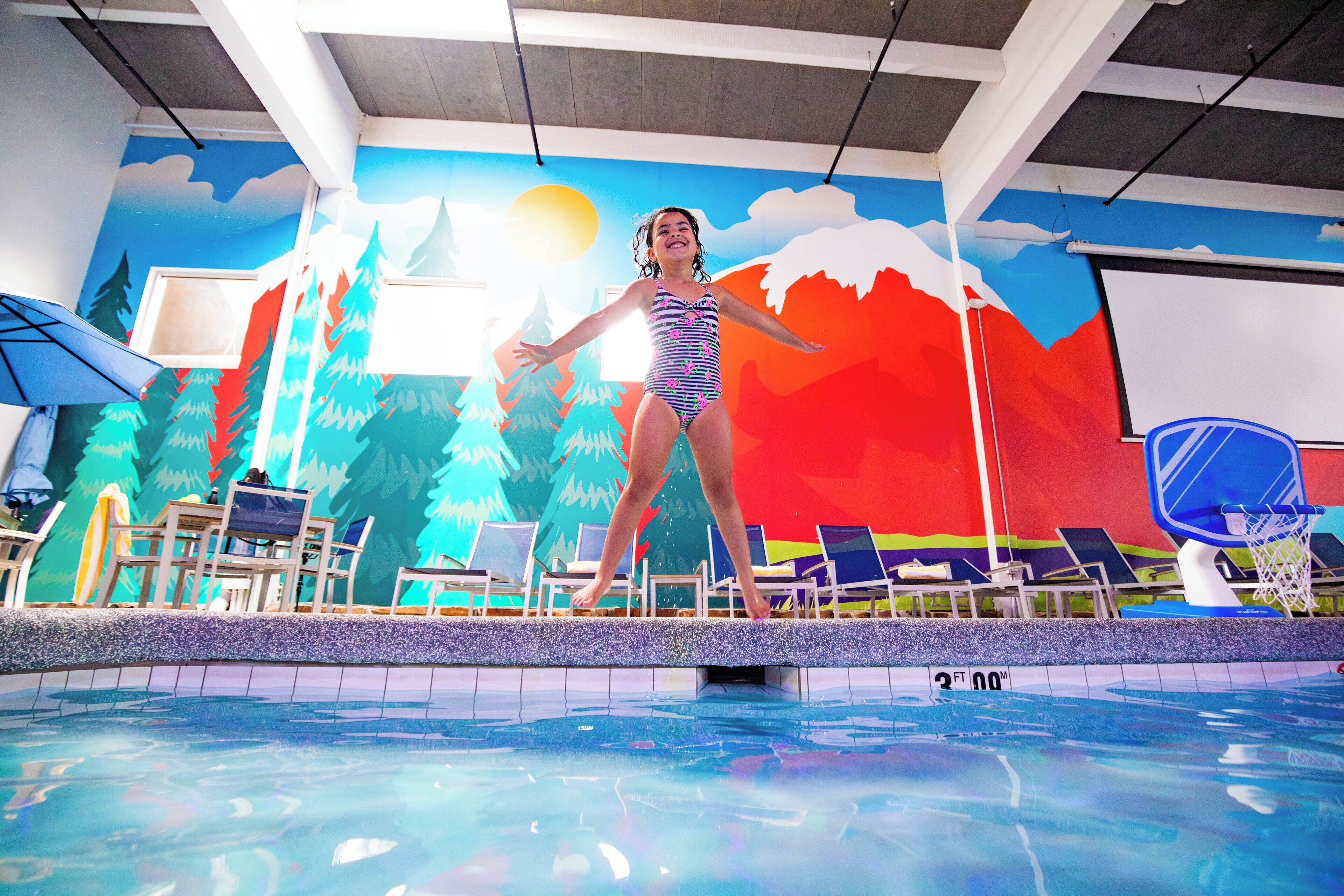 Une piscine ornée d’une fresque représentant des montagnes et des arbres, avec des chaises longues et un panier de basketball.