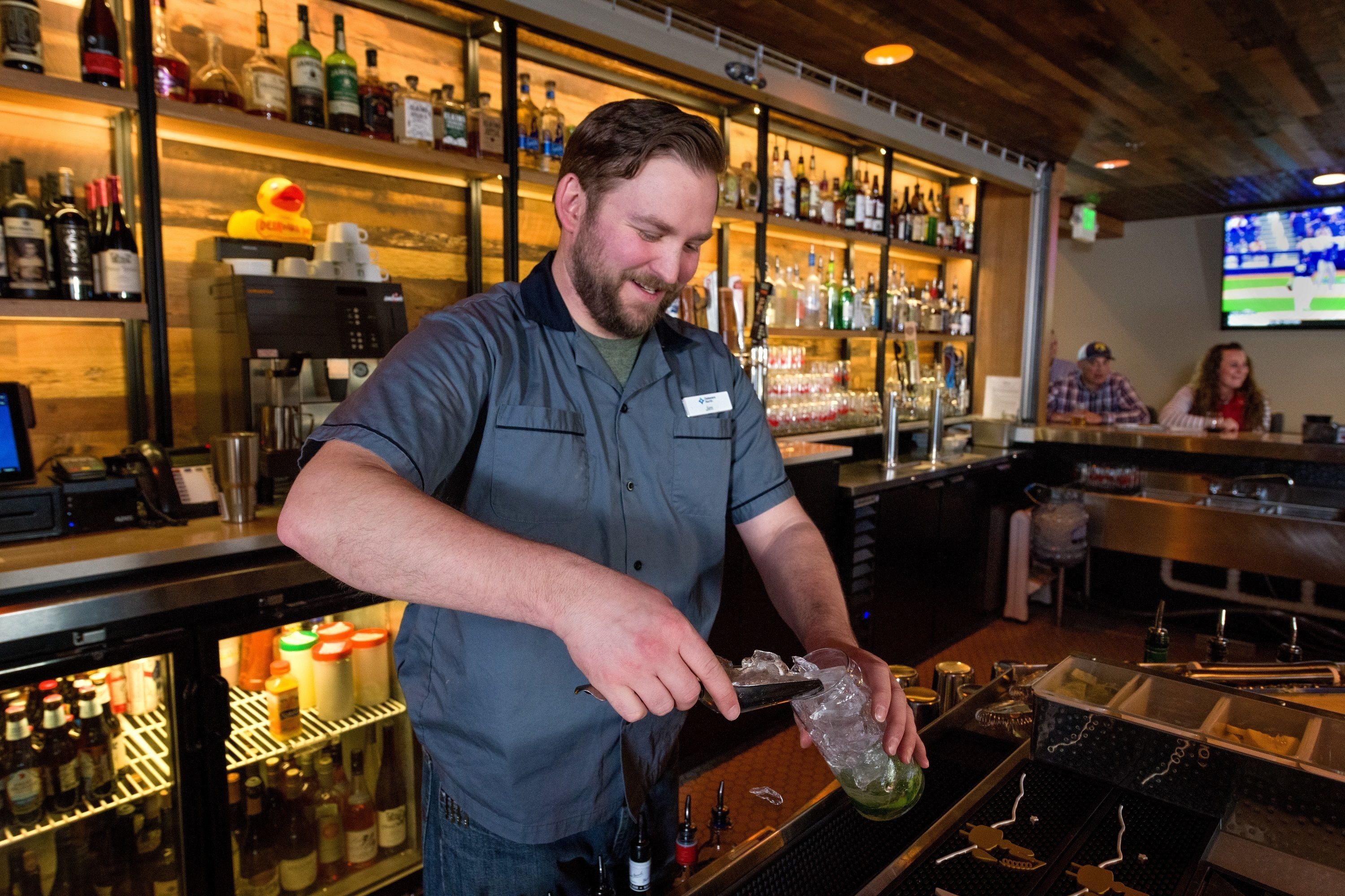 Un barman preparando una bebida detrás de la barra, con estantes llenos de botellas y una máquina de café.