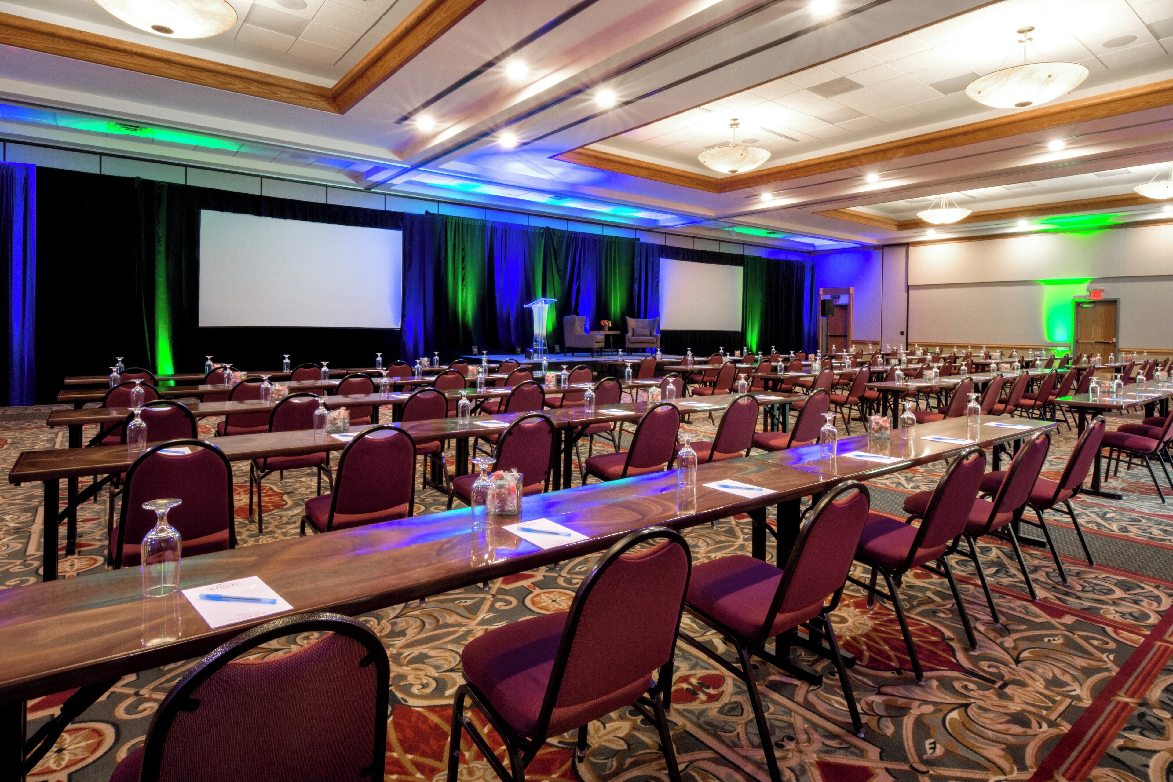 A conference room with rows of tables and chairs, a stage with a screen, and a carpeted floor.