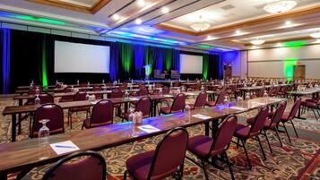 A conference room with rows of tables and chairs, a stage with a screen, and a carpeted floor.