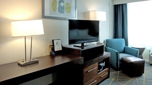 A hotel room with a flat-screen TV, a desk with a lamp, a blue armchair, and a small ottoman.