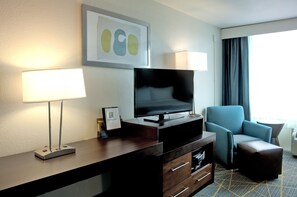 Quarto de hotel com televisão de tela plana, escrivaninha com abajur, poltrona azul e um pequeno banquinho.