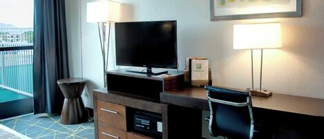 A hotel room with a bed, a desk with a flat-screen TV, a chair, a desk lamp, and a window with a view of a building.