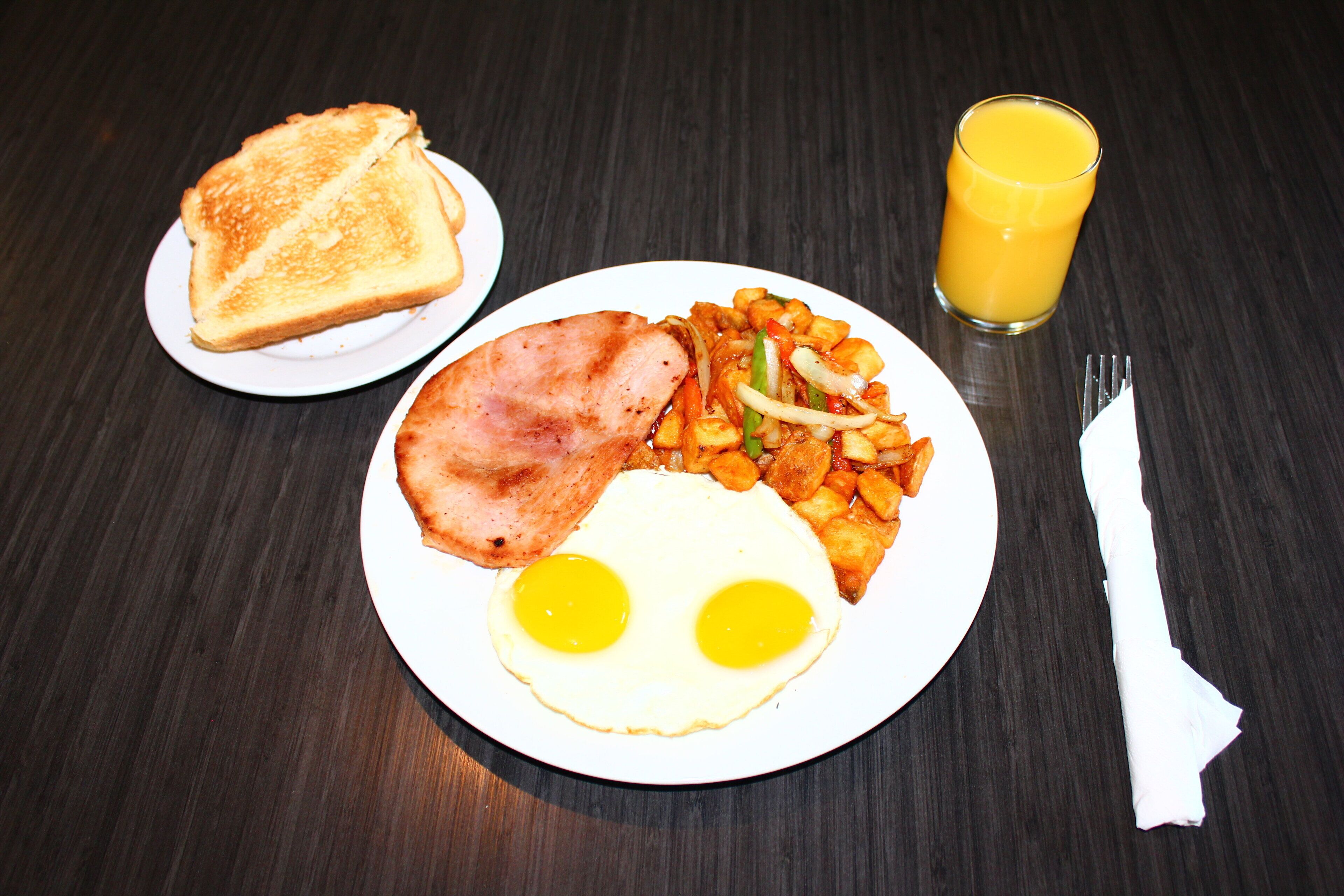 A plate with eggs, ham, and potatoes, a glass of orange juice, and a slice of toast on a dark wooden table.