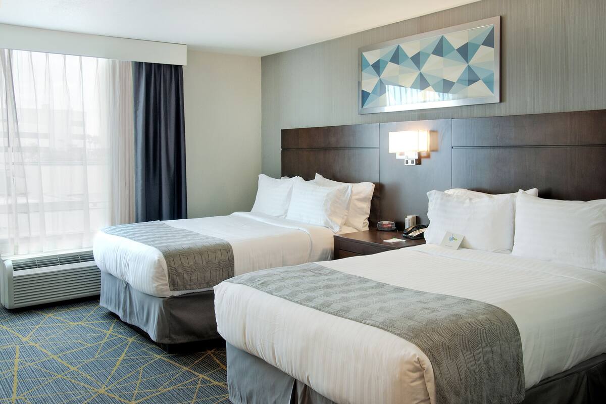 A hotel room with two beds, a large window with curtains, a wall-mounted artwork, and a nightstand with a lamp.
