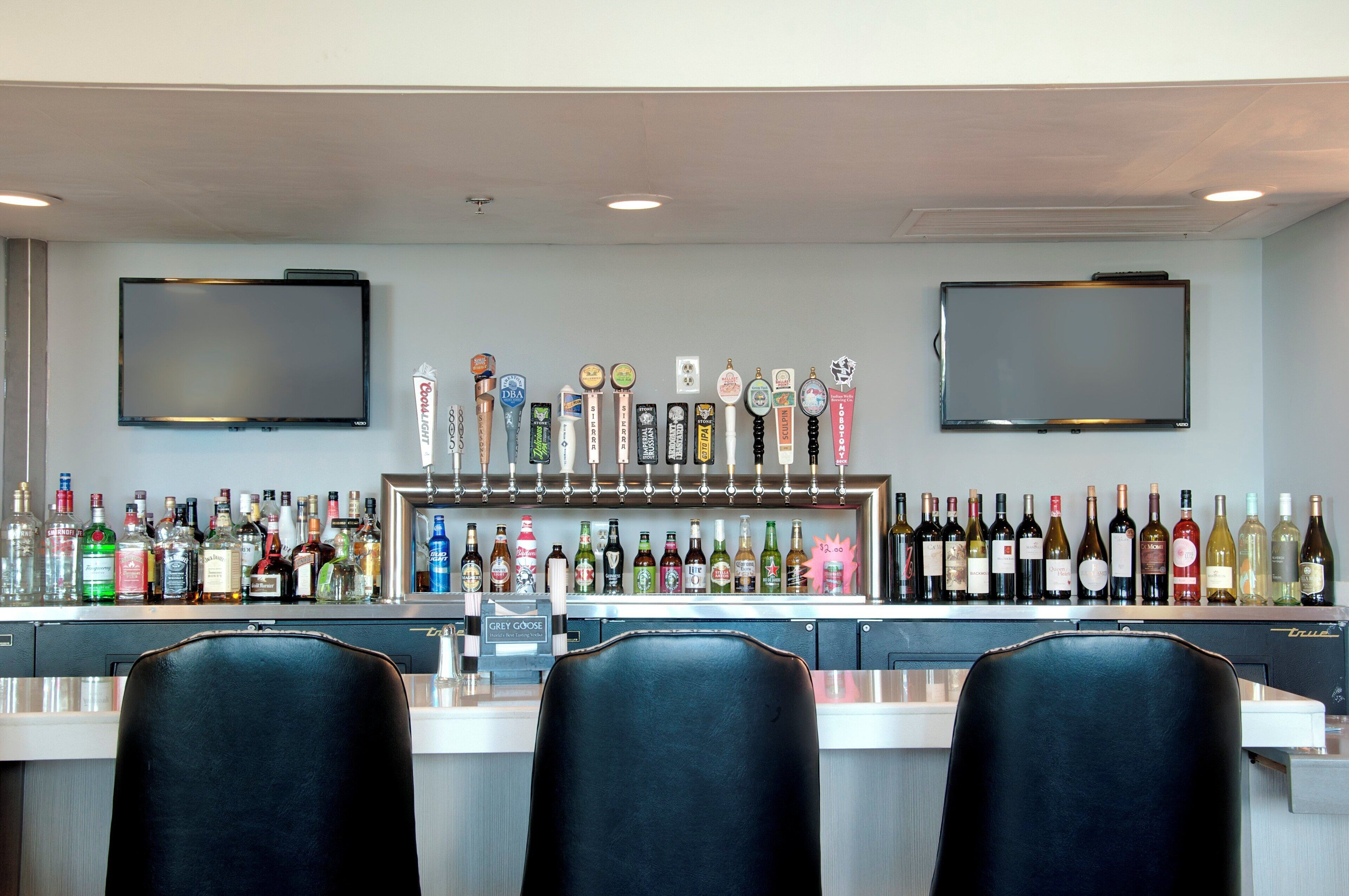 A bar area with multiple taps, a selection of liquor bottles, and two televisions mounted on the wall.