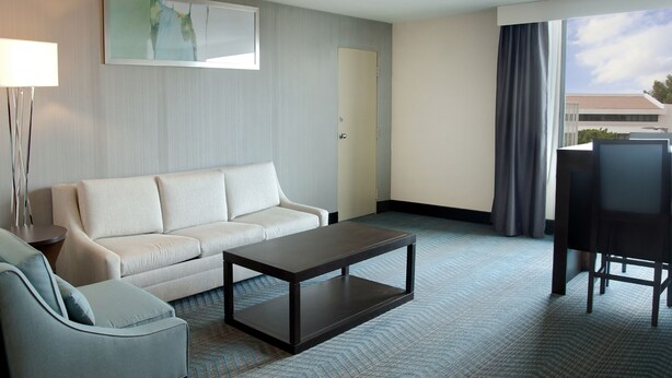 A hotel room with a sofa, a coffee table, a chair, a lamp, a painting, and a view of the outside.