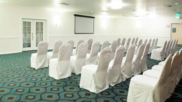 Un salón de conferencias con filas de sillas cubiertas con fundas blancas, una alfombra con un diseño geométrico y una puerta con un panel de vidrio.