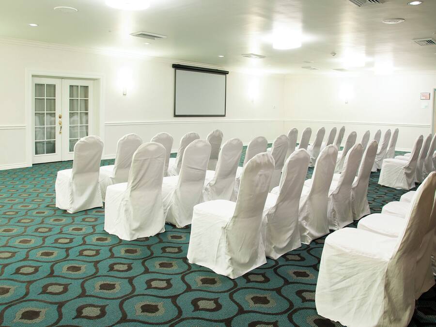 A conference room with rows of chairs covered in white chair covers, a carpet with a geometric pattern, and a door with a glass panel.