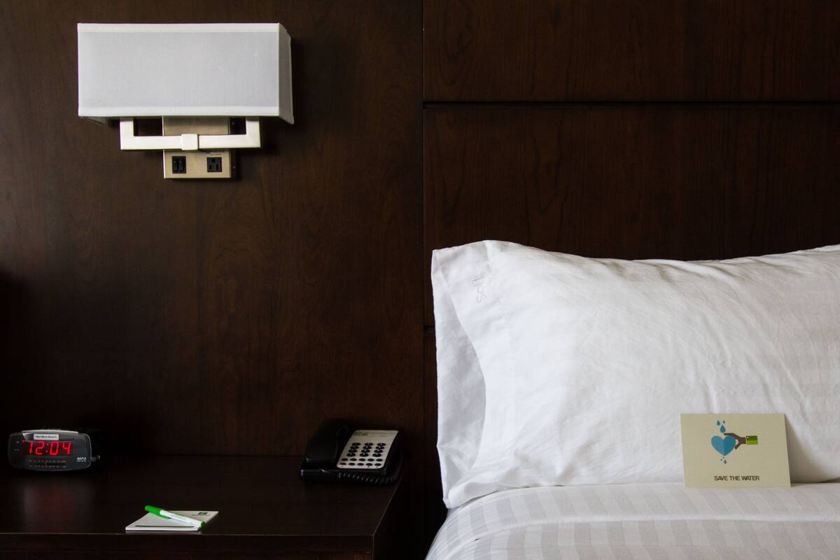 A hotel room with a wooden headboard, a bedside table with a clock, a remote control, a notepad, and a pillow with a 'Save the Water' card.