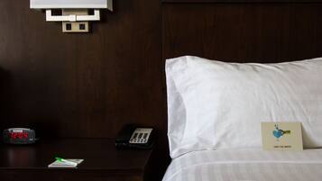 A hotel room with a wooden headboard, a bedside table with a clock, a remote control, a notepad, and a pillow with a 'Save the Water' card.