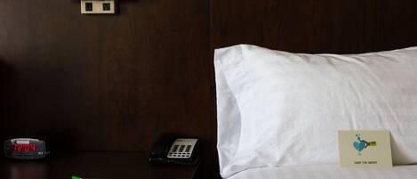 A hotel room with a wooden headboard, a bedside table with a clock, a remote control, a notepad, and a pillow with a 'Save the Water' card.