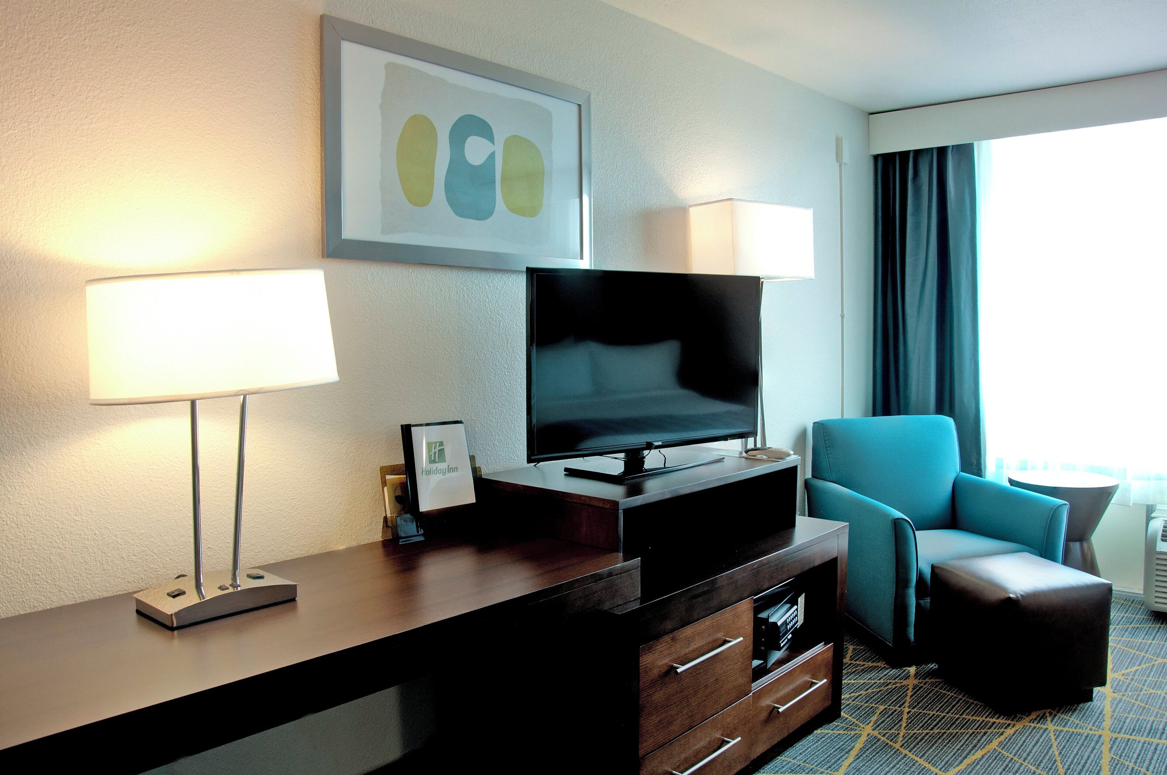 A hotel room with a flat-screen TV on a wooden dresser, a blue armchair, a lamp, and a framed artwork on the wall.