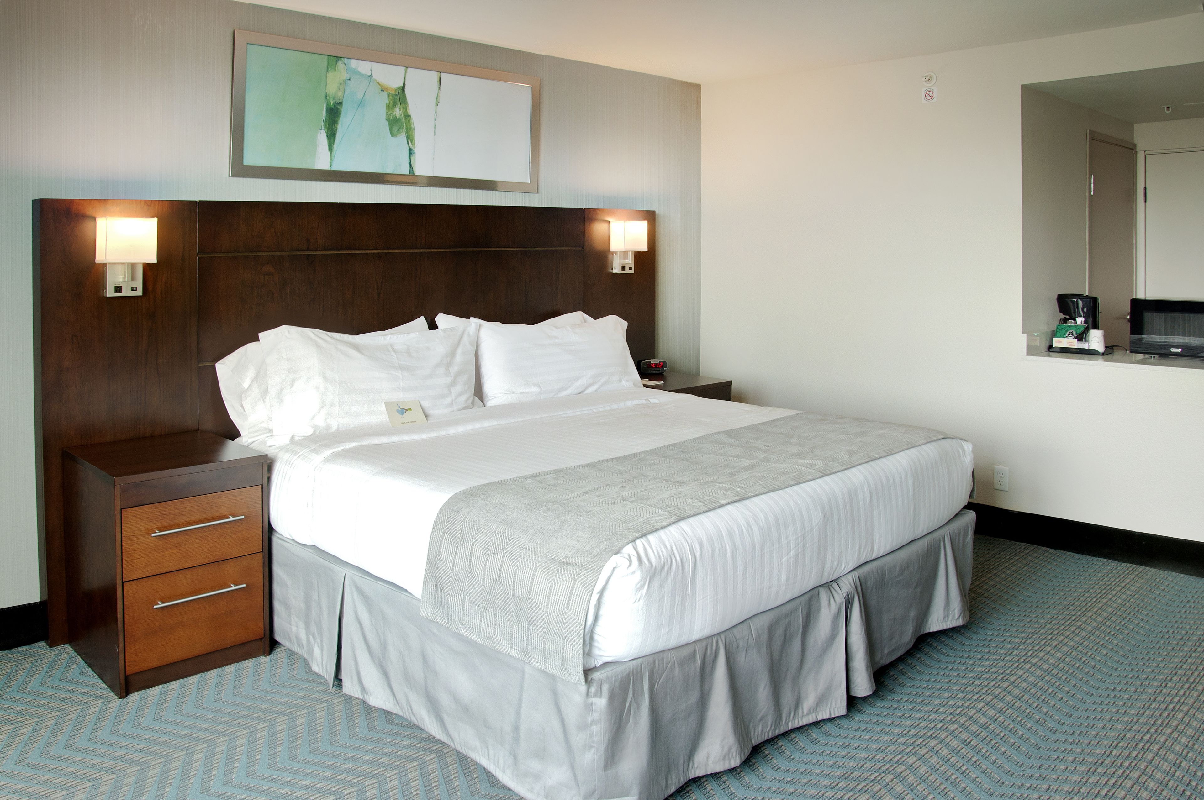 A hotel room with a large bed, a wooden nightstand with drawers, a wall-mounted lamp, and a painting above the bed.