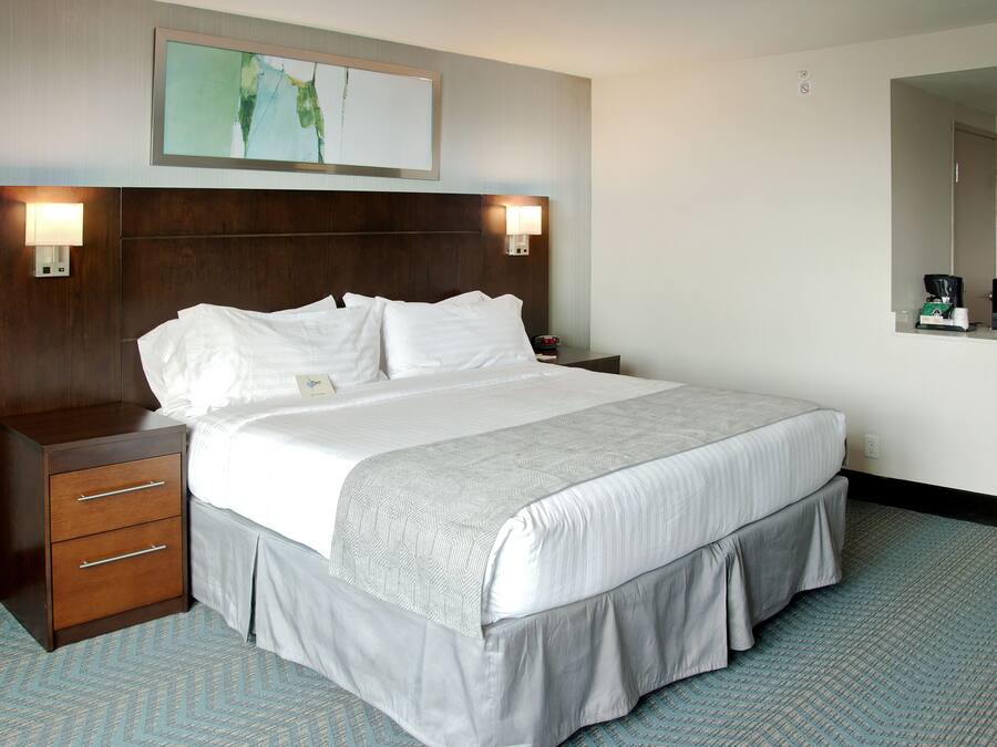 A hotel room with a large bed, a wooden nightstand with drawers, a wall-mounted lamp, and a painting above the bed.
