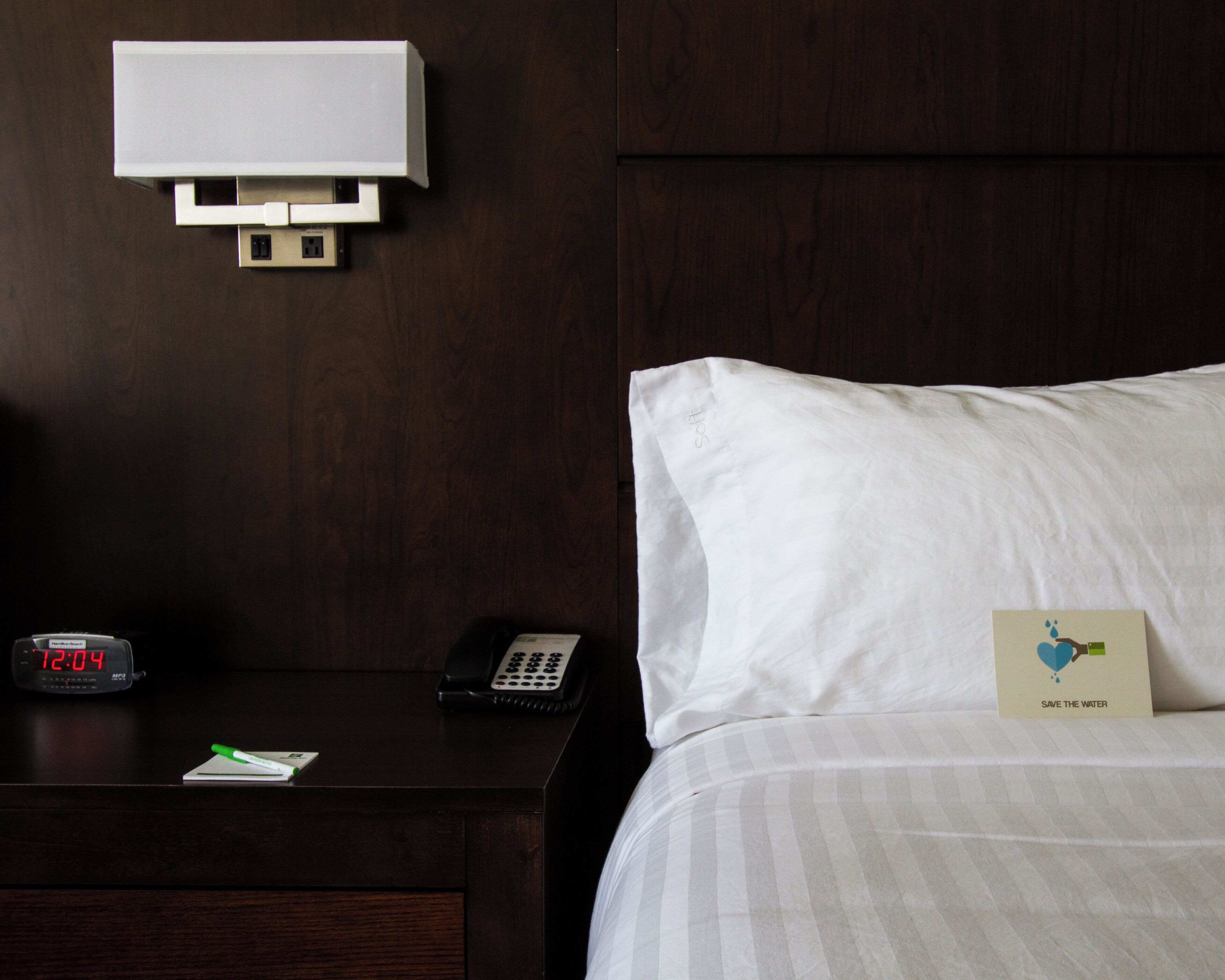 A hotel room with a bed, a bedside table with a clock, a phone, and a card on the pillow.