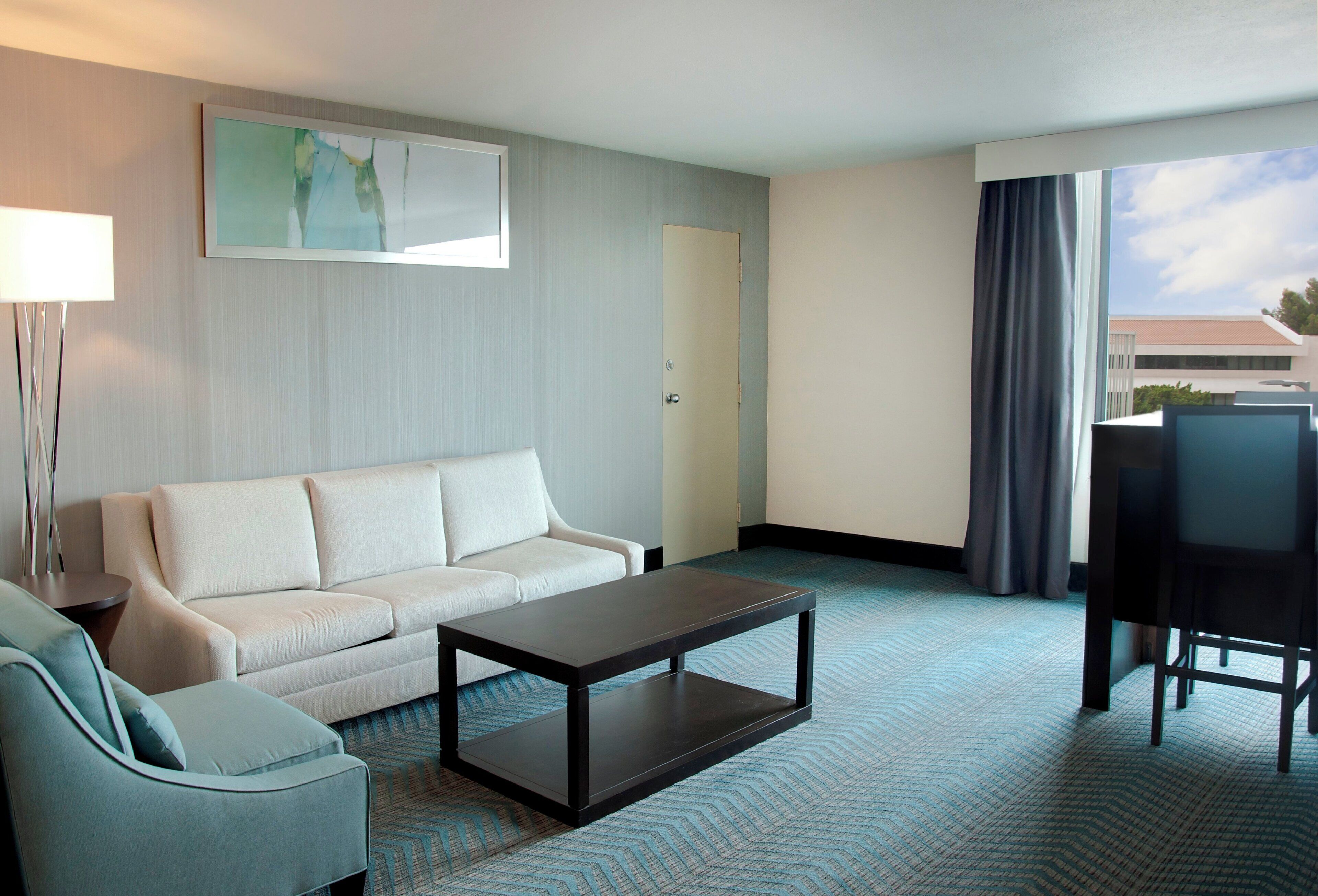 A hotel room with a sofa, a small table, a chair, a lamp, a painting, a door, and a window with curtains.