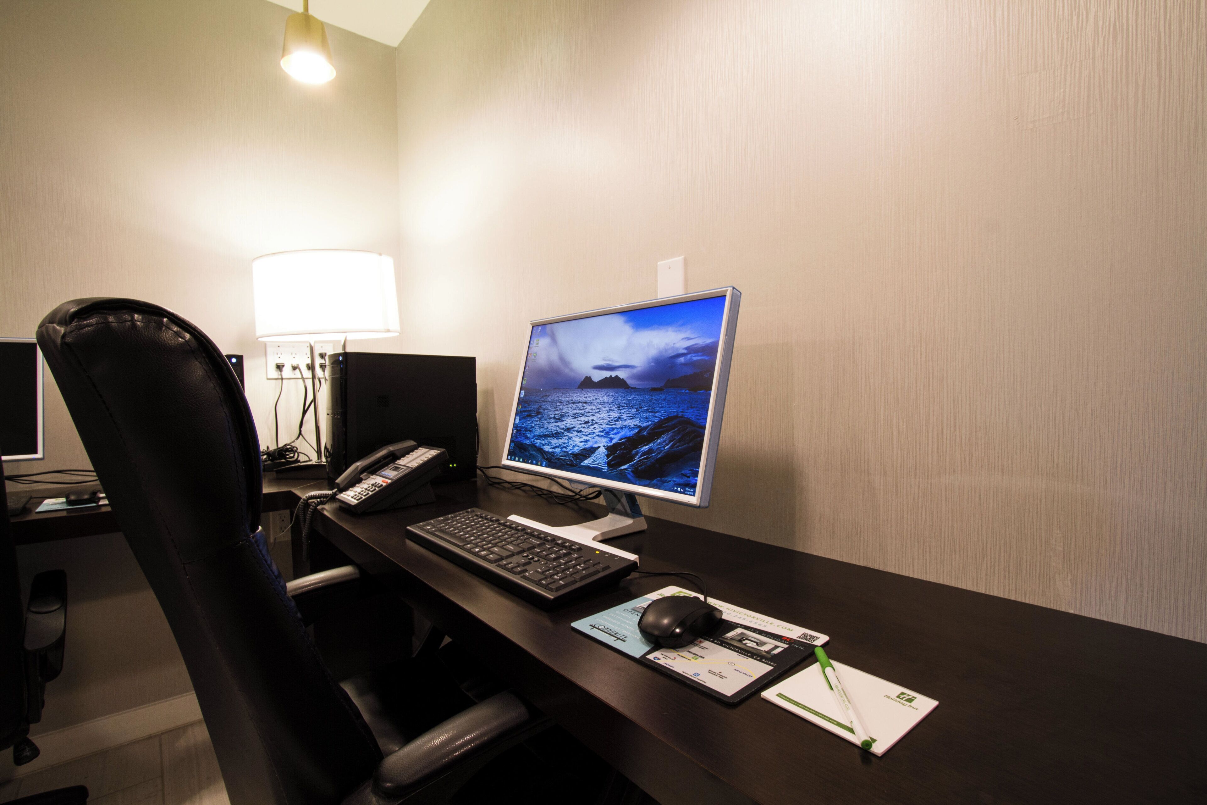 A hotel room with a desk, computer, chair, lamp, and a view of the sea on the monitor.