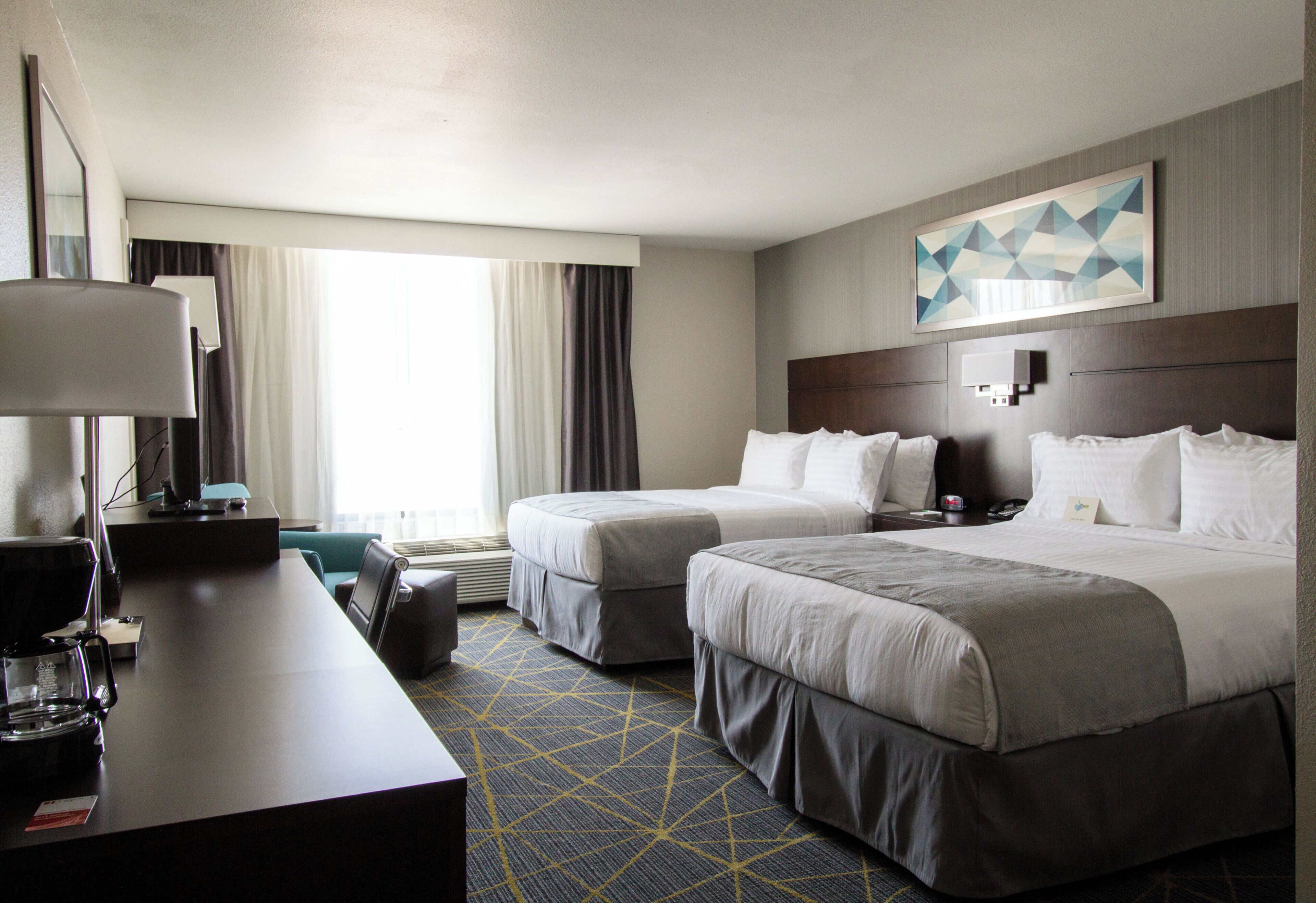 A hotel room with two beds, a desk with a lamp, a coffee maker, and a large window with curtains.