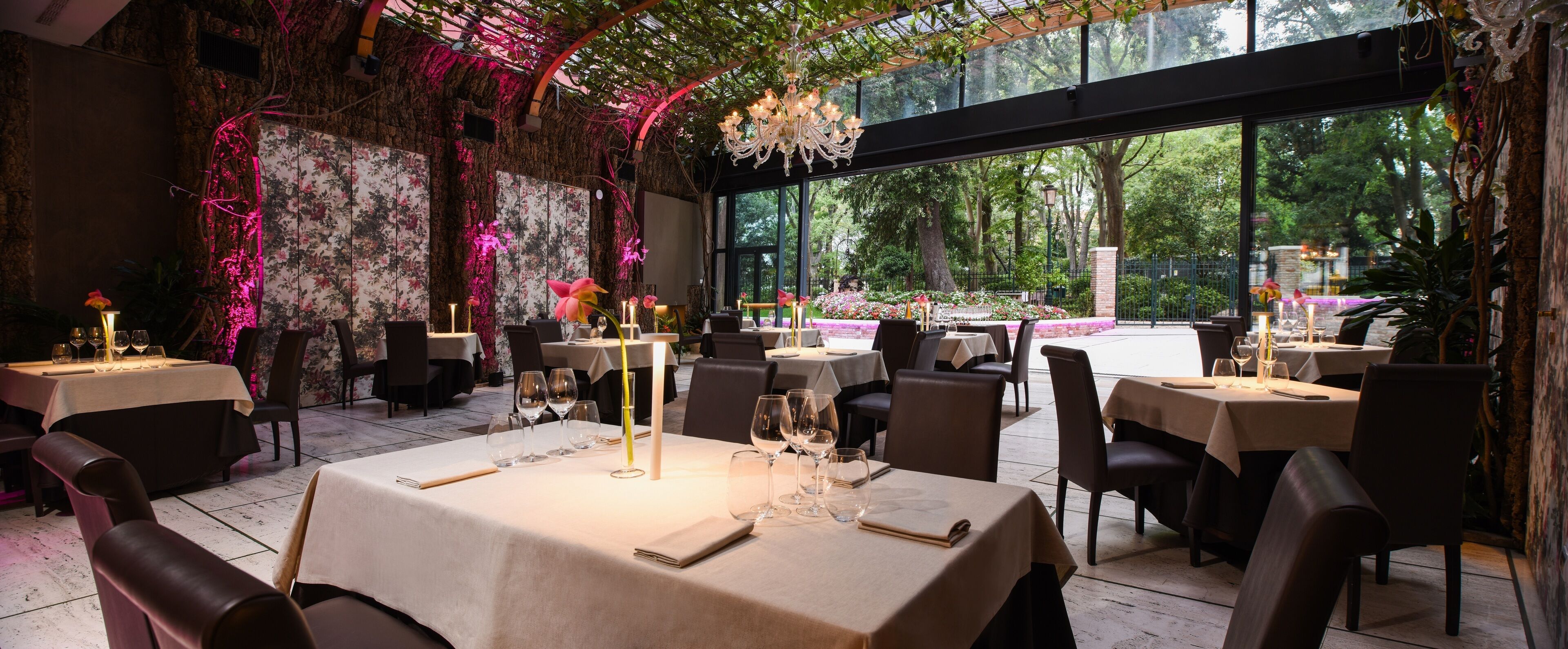 A restaurant with tables set for dining, a chandelier, and a view of trees through large windows.