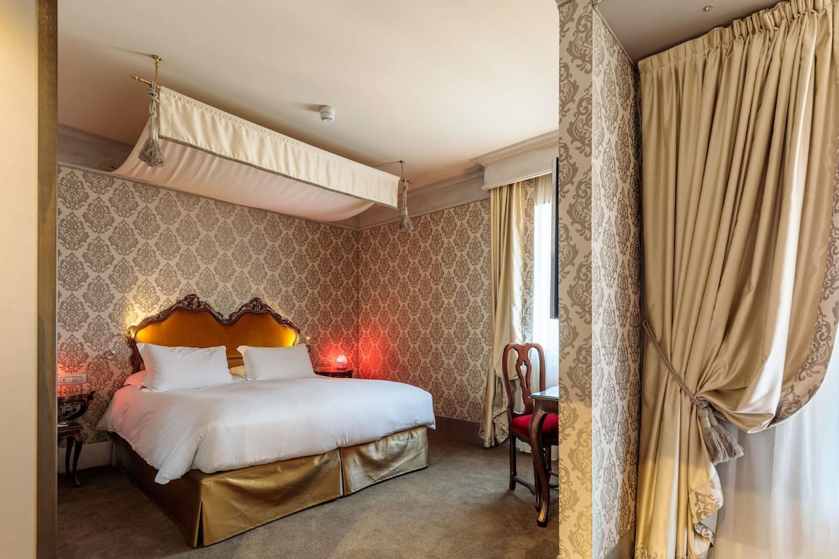 A hotel room with a bed, a nightstand, a chair, a window with curtains, and a patterned wallpaper.