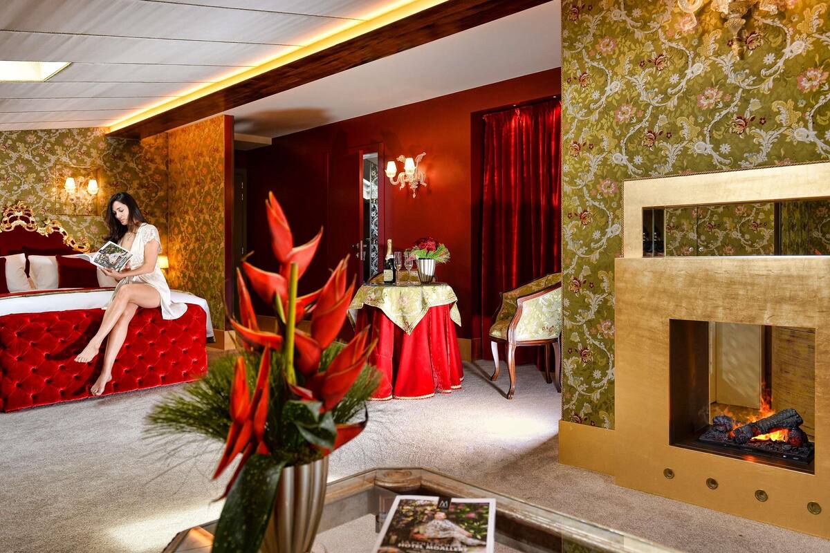 A room with a red upholstered bed, a fireplace, a table with a vase of flowers, and a decorative mirror.