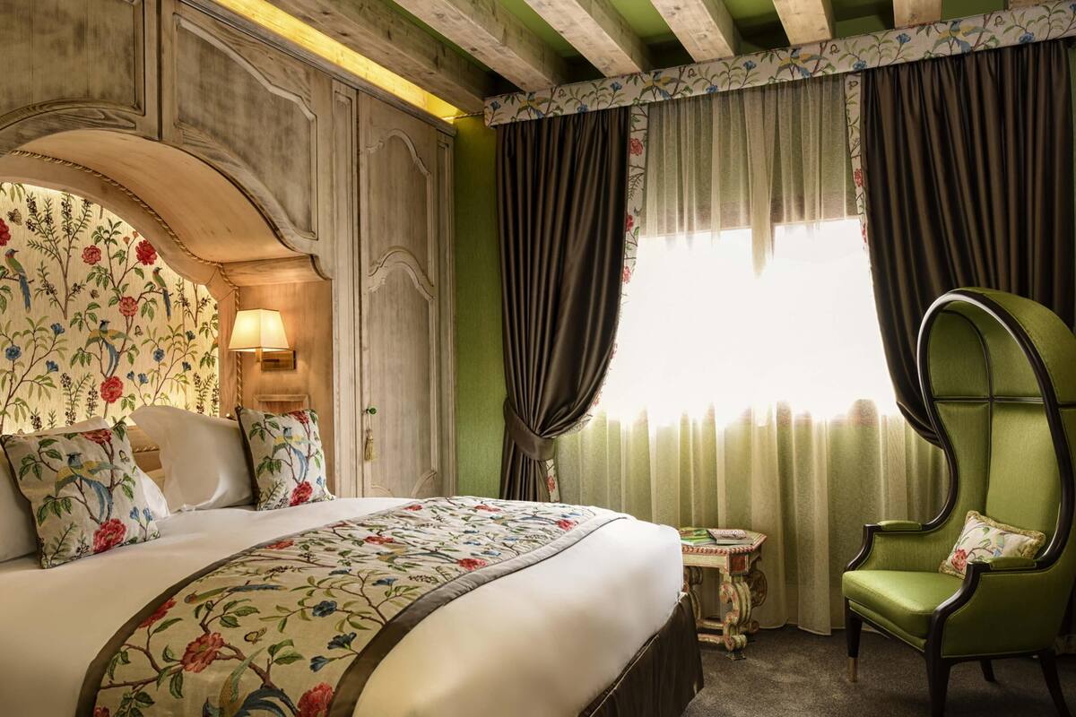 A hotel room with a large bed, a green armchair, a small table, and a window with curtains.