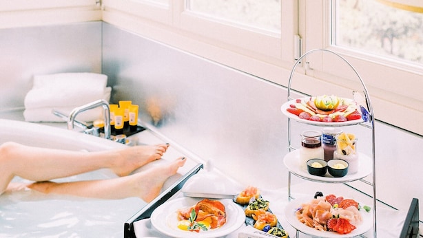 Un entorno de spa con una bañera de hidromasaje, un carrito de servicio de tres niveles con comida y bebidas, y vistas al exterior a través de grandes ventanales.