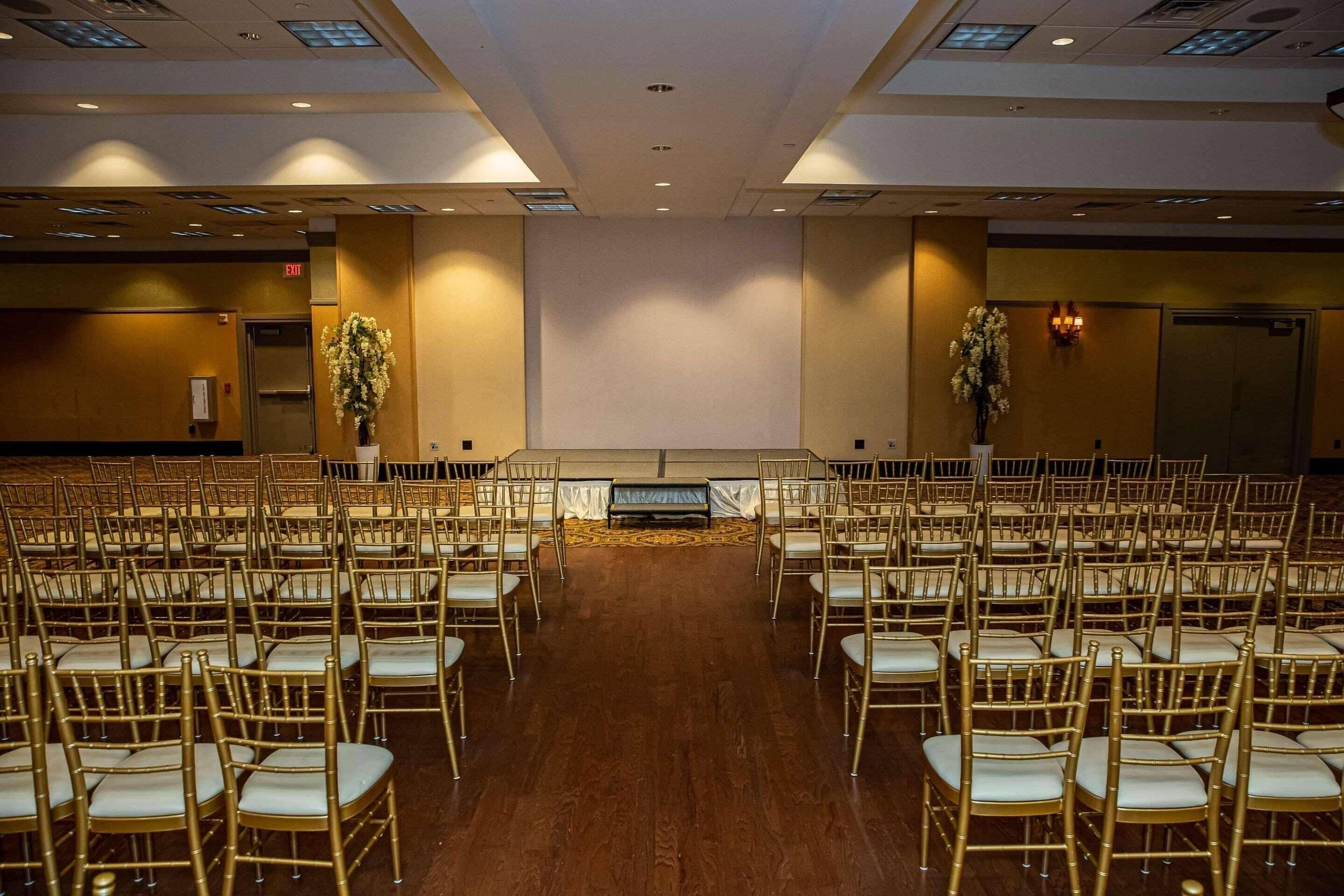 A conference room with rows of golden chairs, a stage at the front, and a large wall with a floral arrangement.