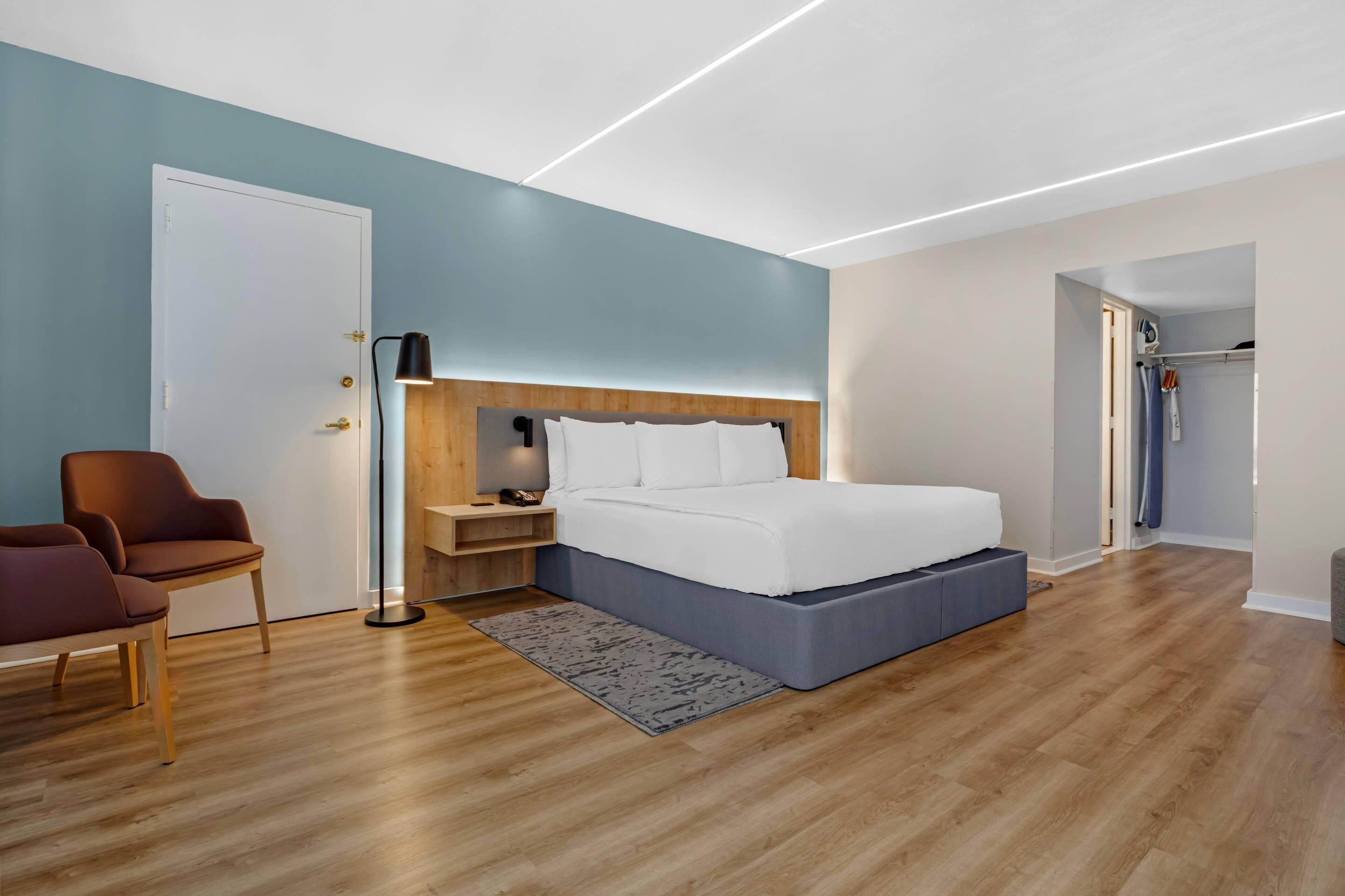 A modern bedroom with a wooden headboard, a bed with white linens, a floor lamp, and a seating area with two chairs.