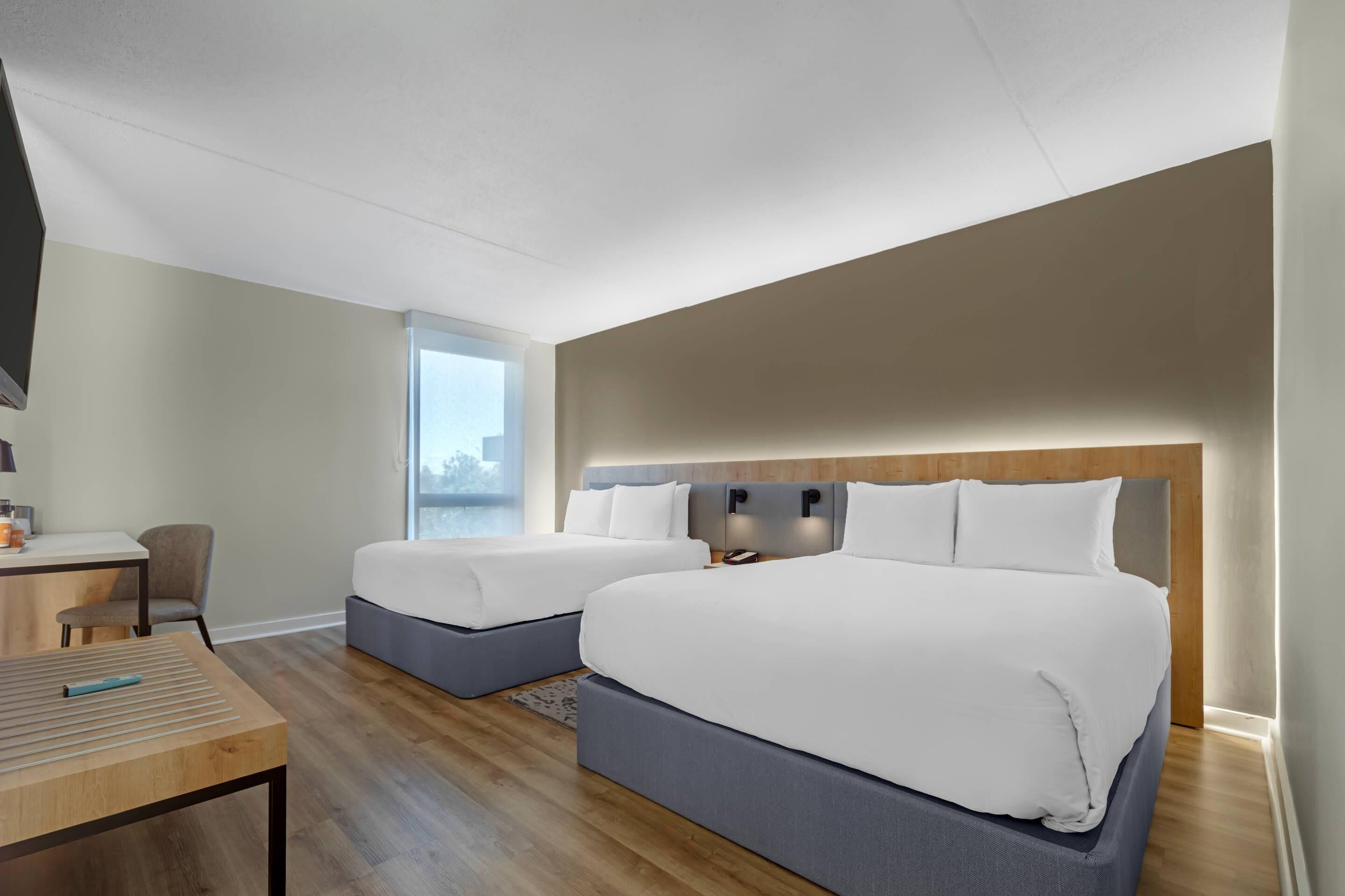 A modern hotel room with two beds, a desk, and a large window offering a city view.