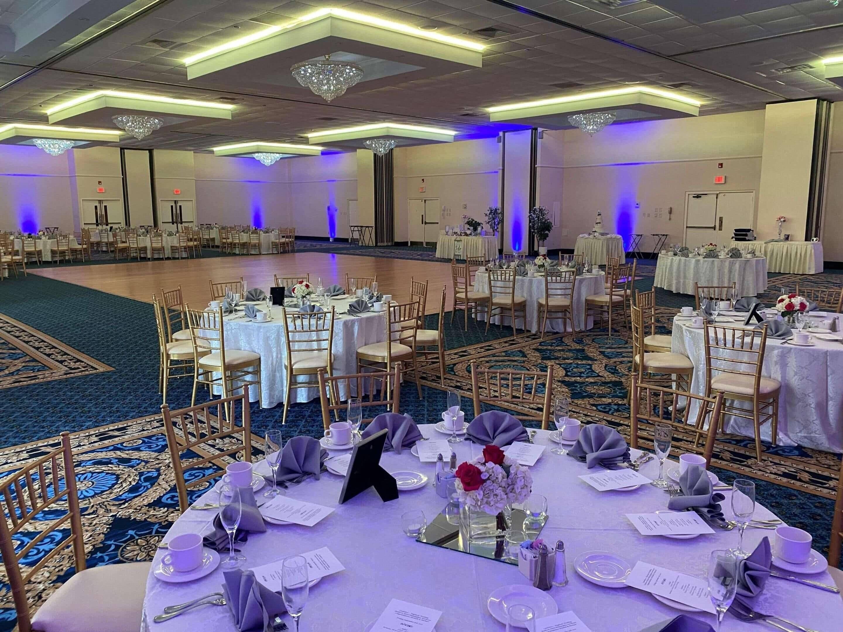Un salón de banquetes con mesas redondas dispuestas para un evento formal, con sillas doradas, manteles blancos y centros de mesa florales.